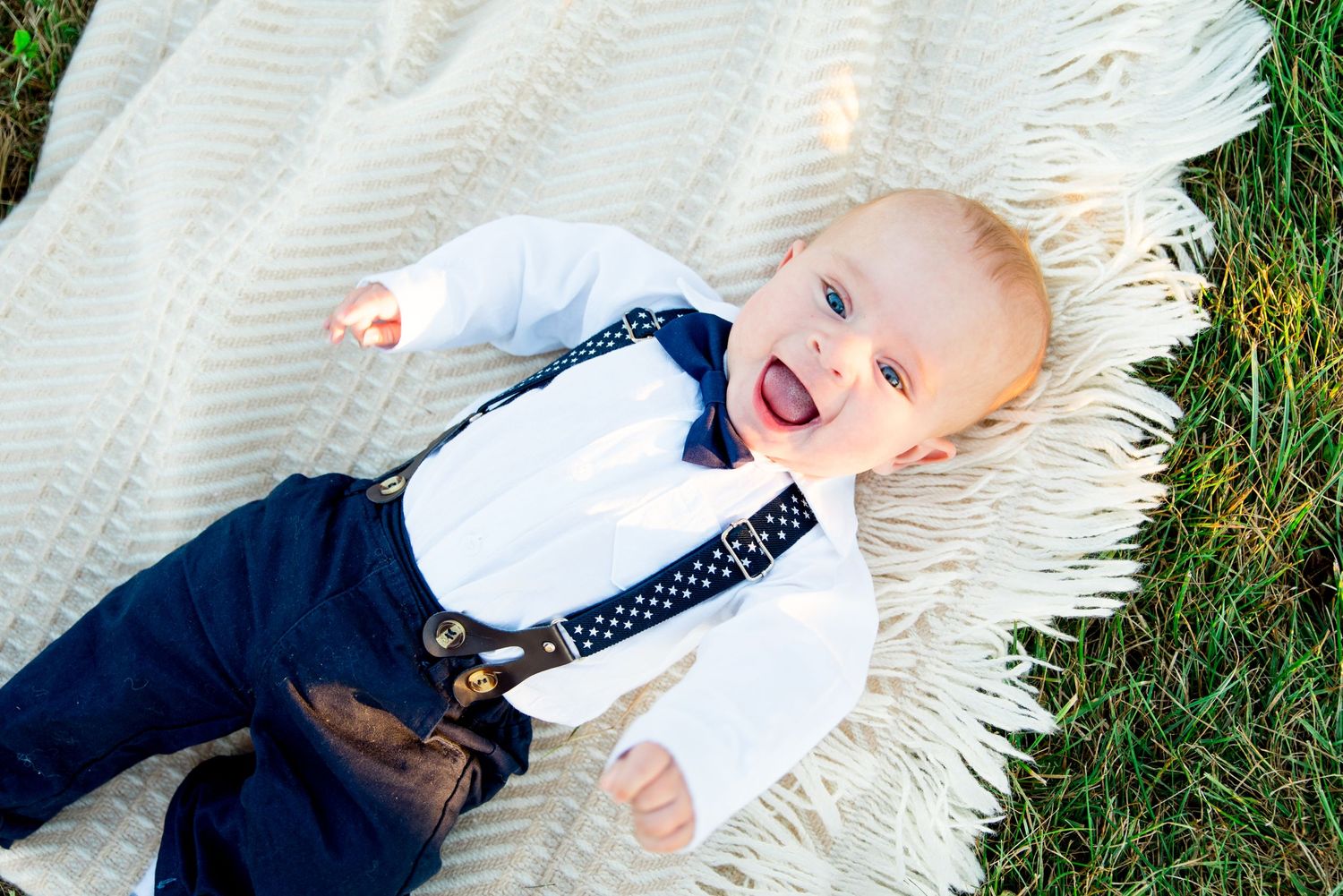 young baby boy in white button down and navy pants and suspenders lays on a blanket and smiles