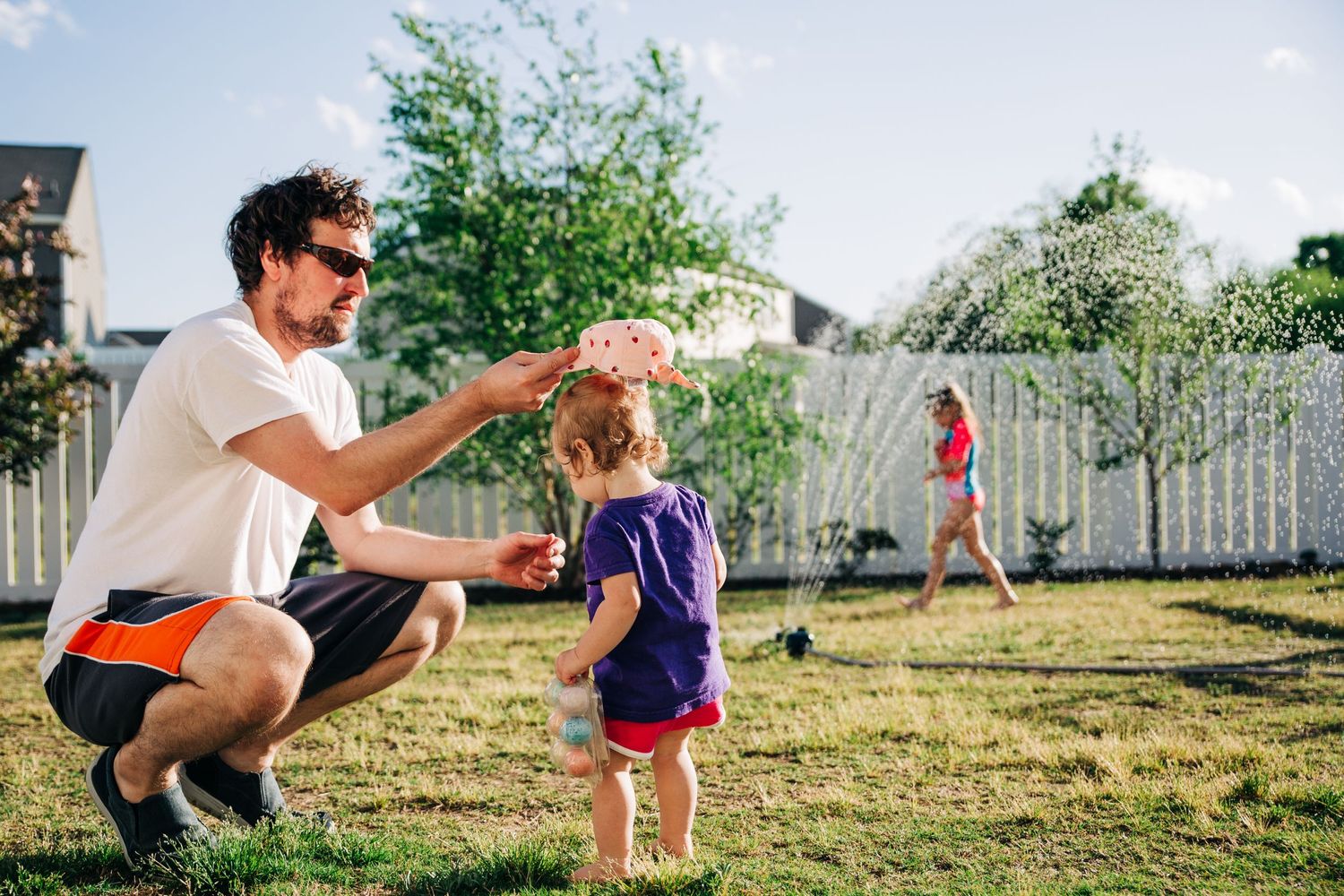 A dad and his two daughters playing outside in the backyard in the sprinkler