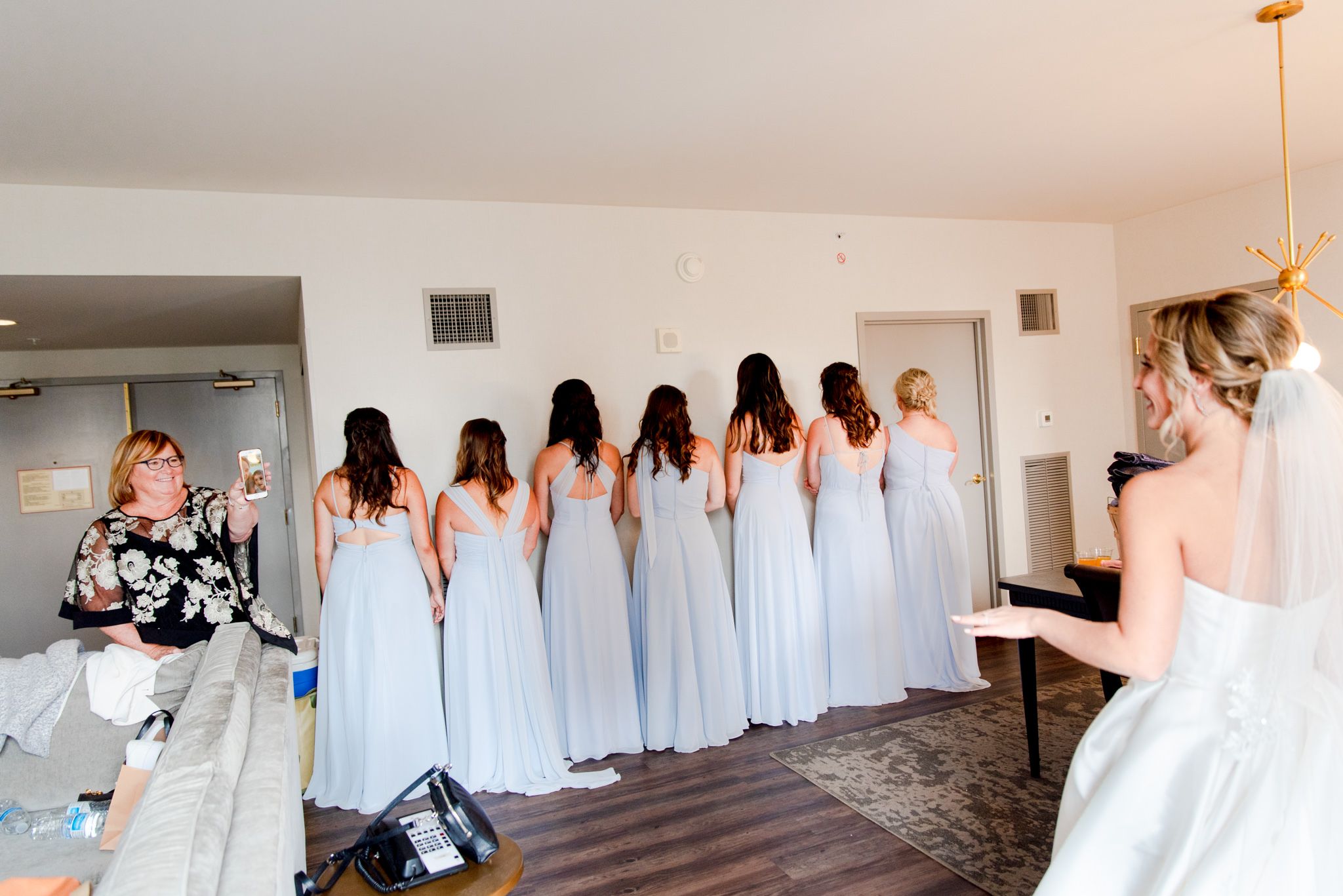 seven bridesmaids in ice blue dresses face the wall and wait to see the bride while mom facetimes family members