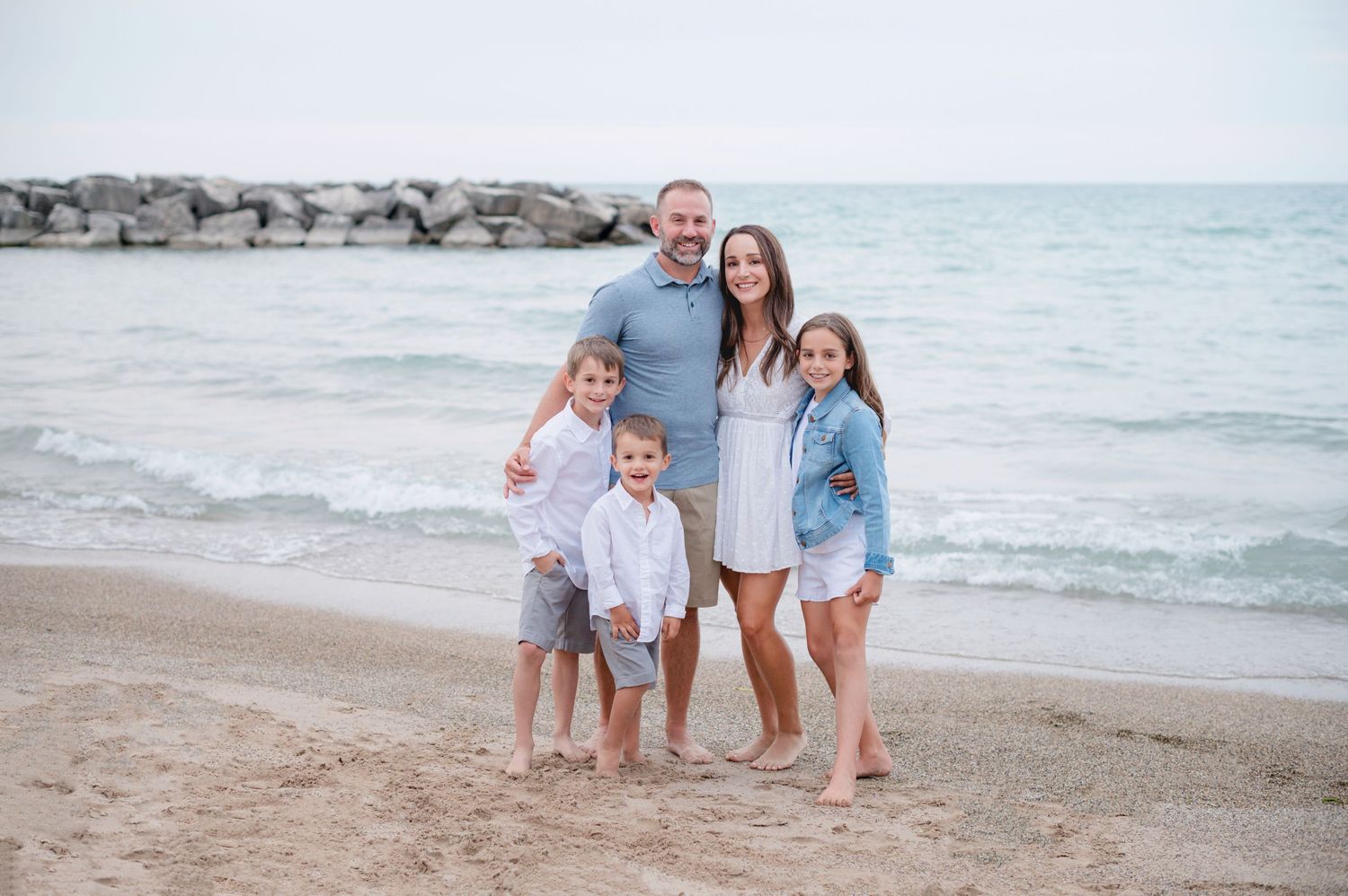 Family poses together on beach with rock jetty and ocean waves in background.