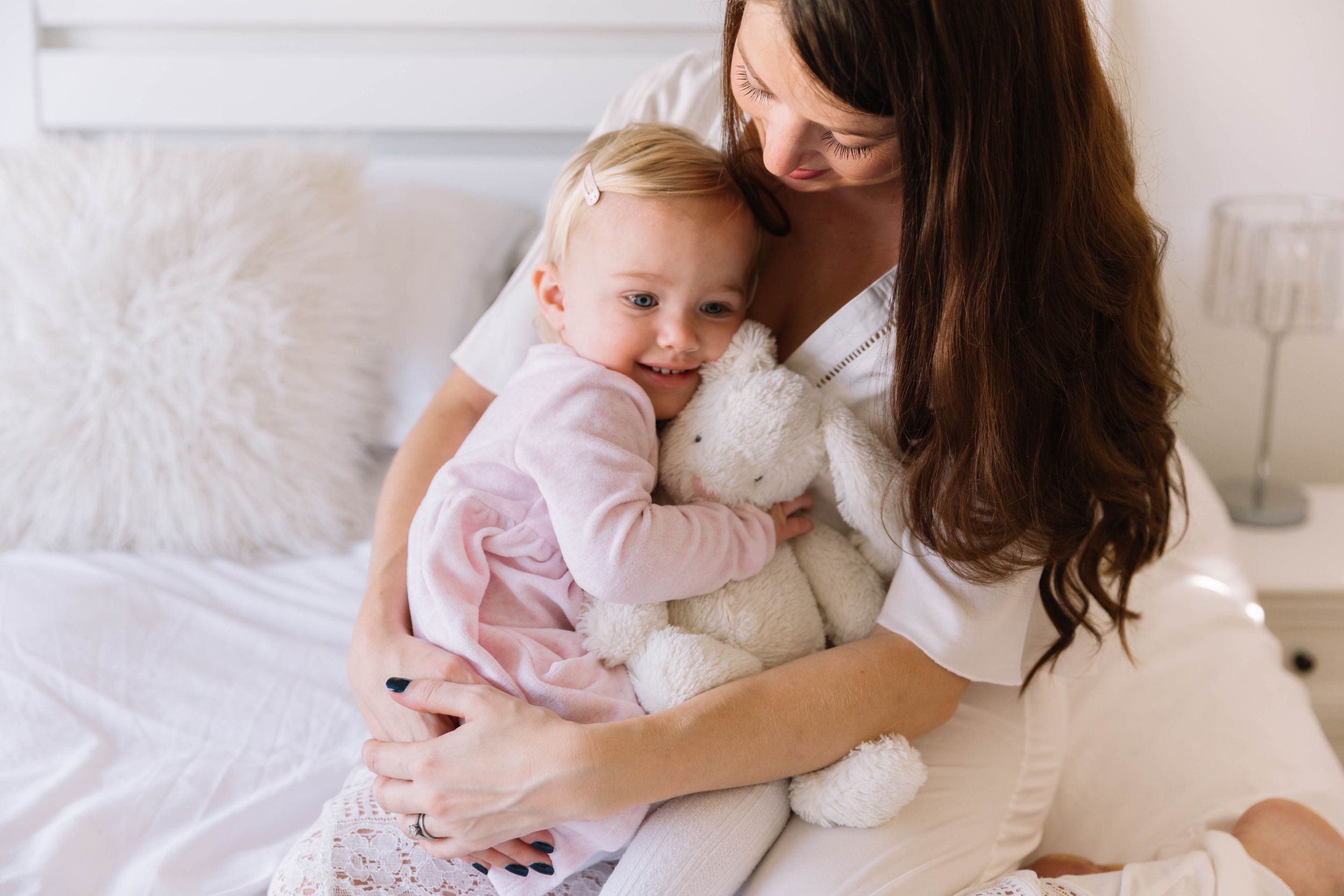 pregnant mum in white dress and toddler daughter in pink dress cuddling on bed