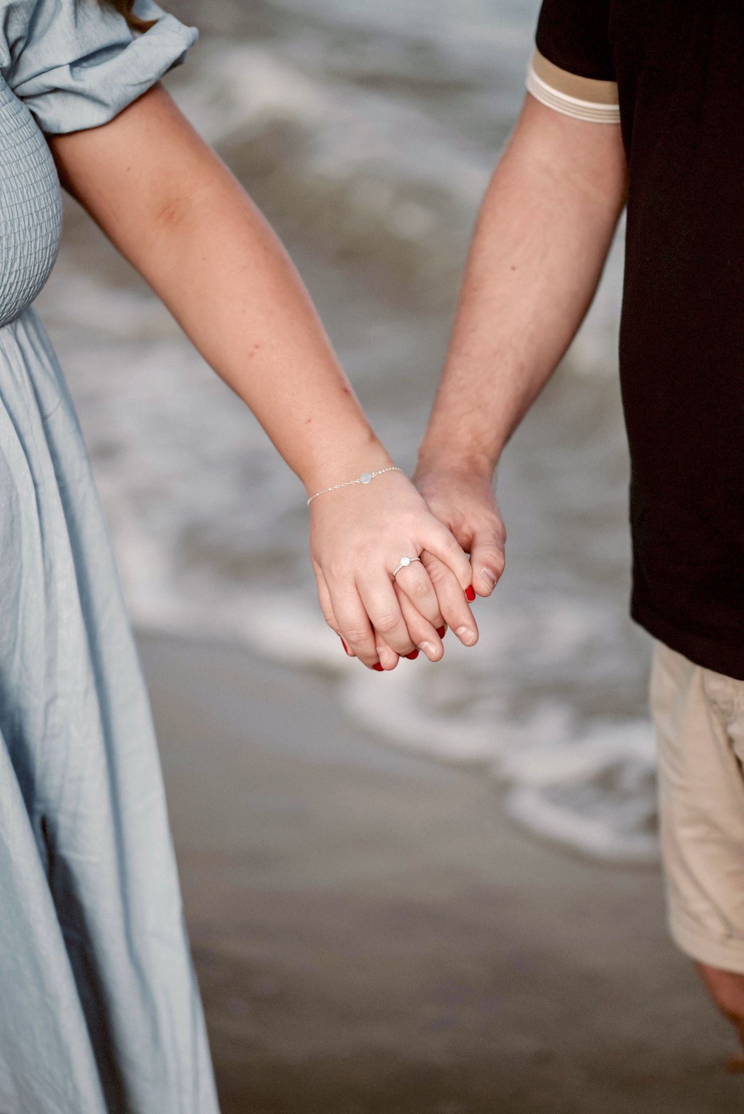 Close-up of two people holding hands tenderly on a beach with ocean waves and light blue dress visible.