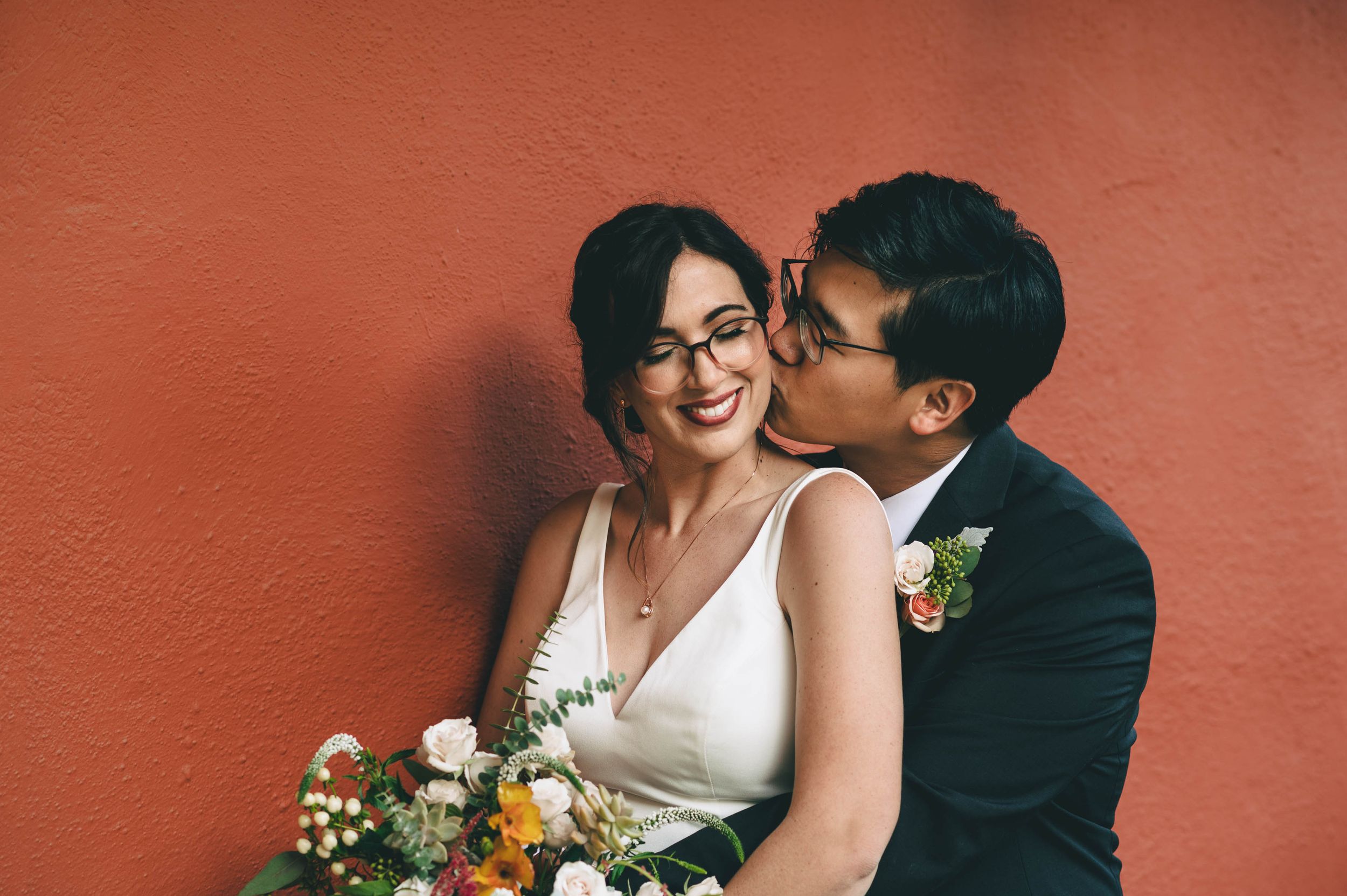 Groom kisses bride on cheek, in front of the red Adobe walls at Cooper Molera in Monterey.