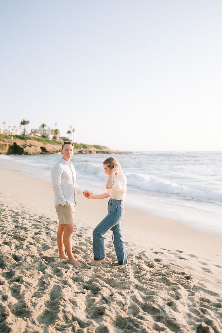 Couple sharing special moment on sandy beach with coastal cliffs and palm trees in distance.