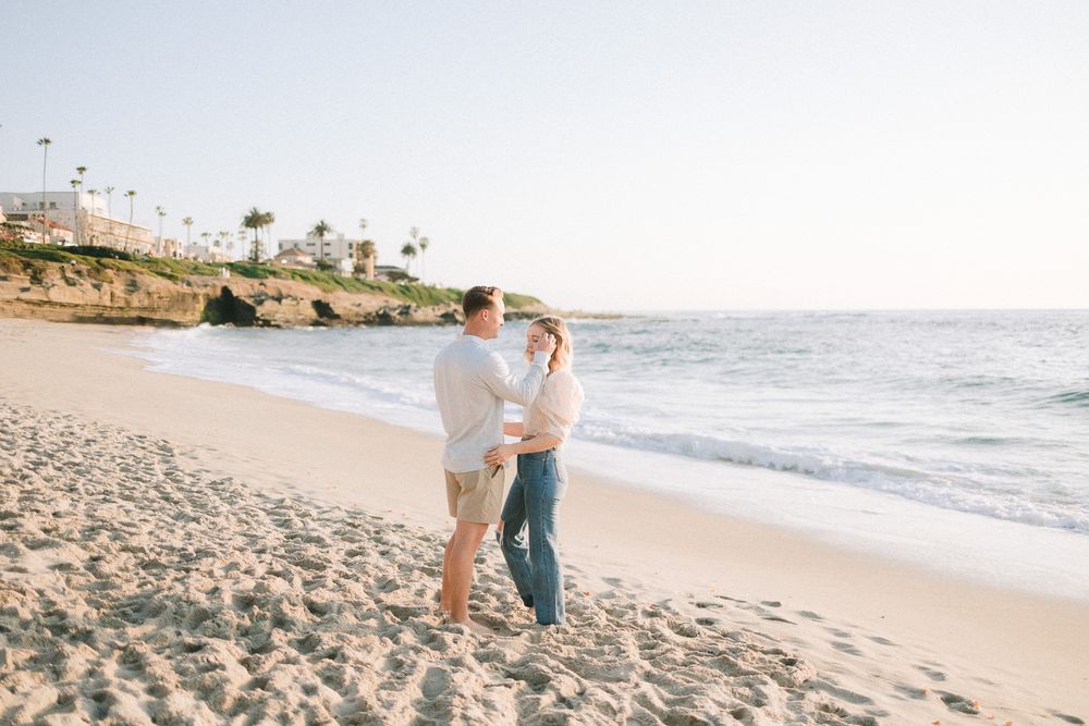 A couple walking together on a scenic beach with cliffs and palm trees in the background during sunset.