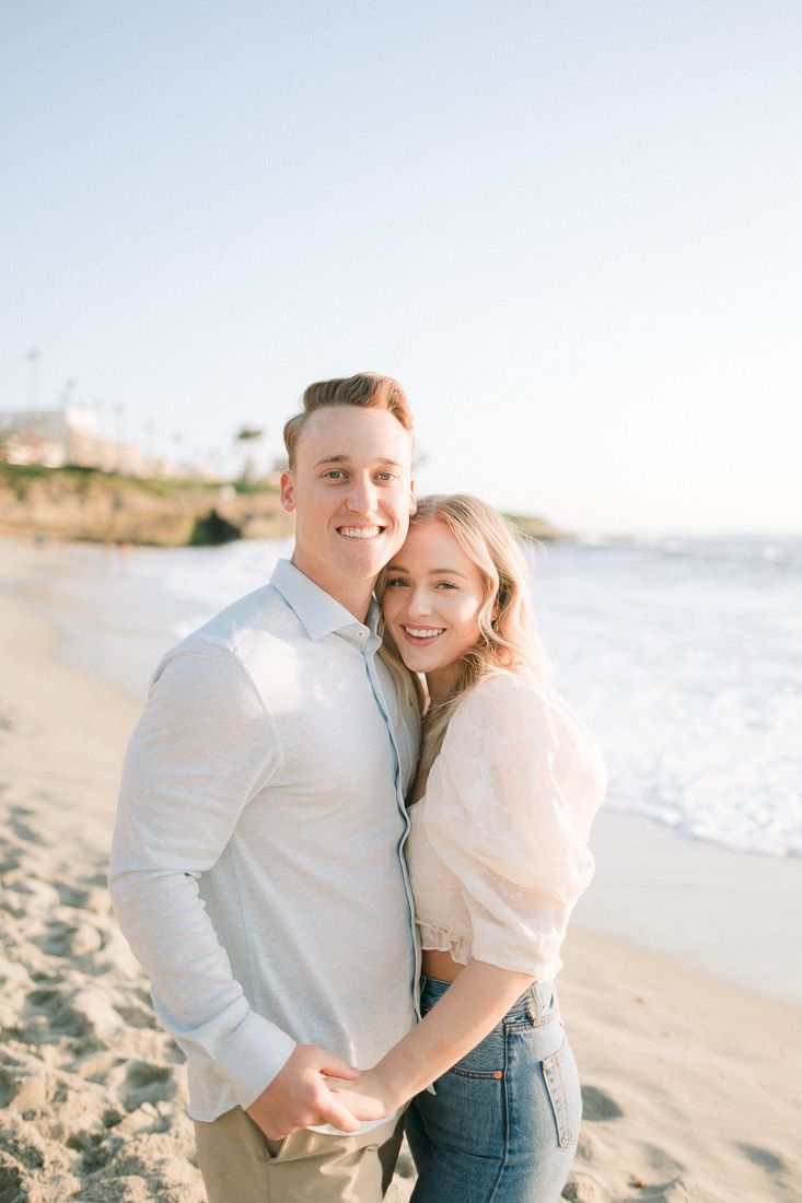 A happy couple smiling together on a sunny beach with ocean waves in the background.