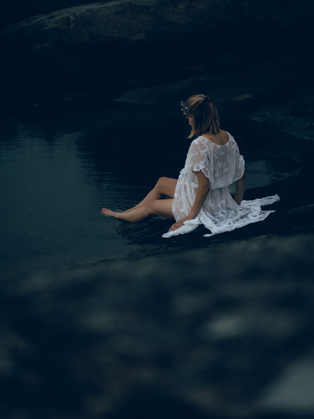 Person in flowing white dress sitting by dark water's edge in moody lighting.