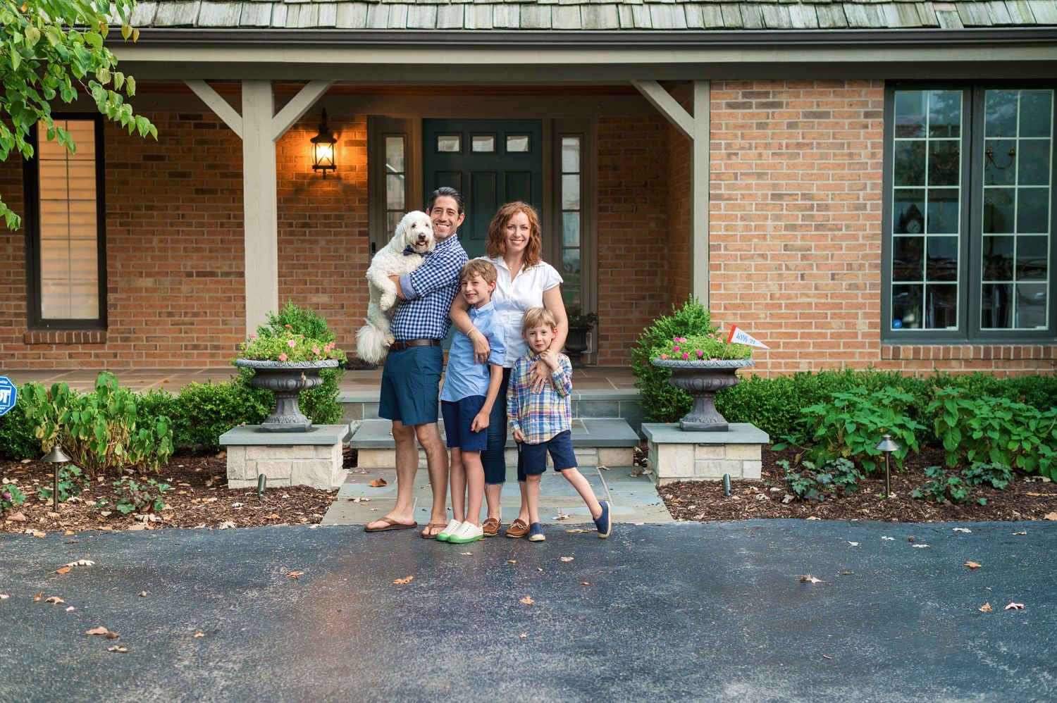 Family and dog pose together on front porch of brick house with planters.