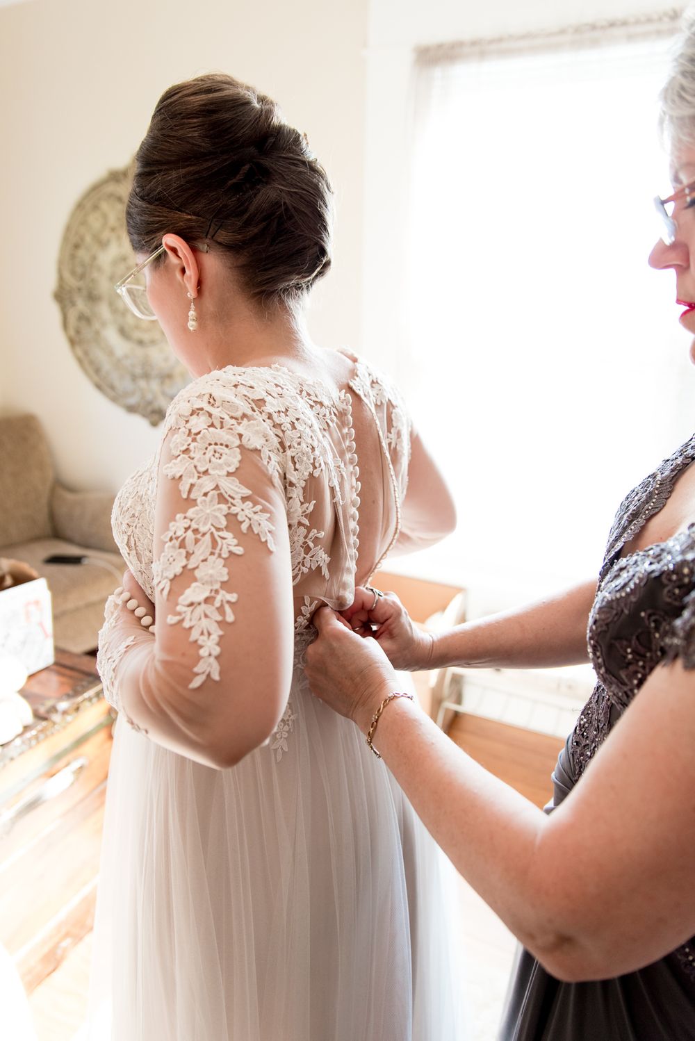 mother of the bride buttoning up bride's wedding dress in an all white room