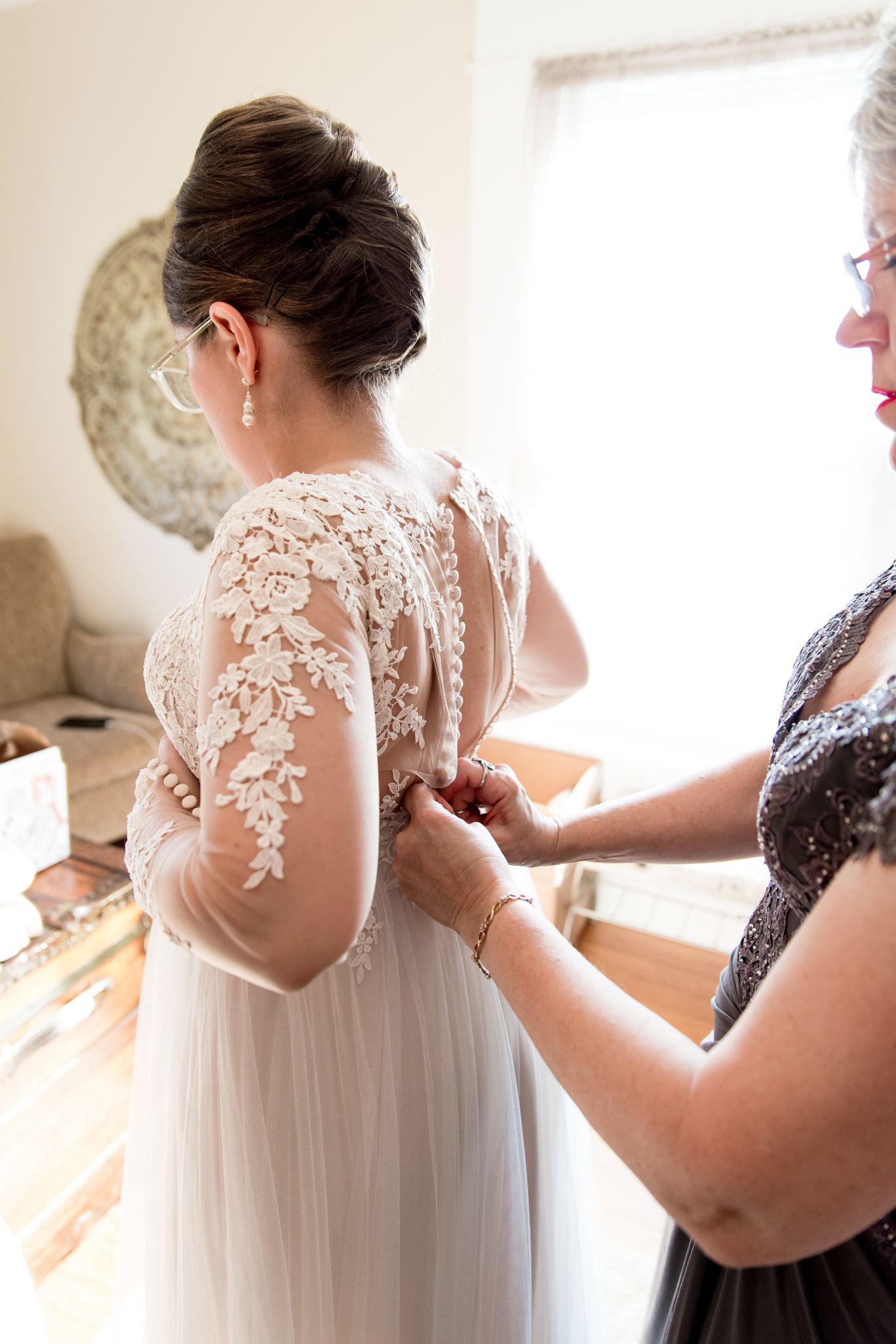 mother of the bride buttoning up bride's wedding dress in an all white room