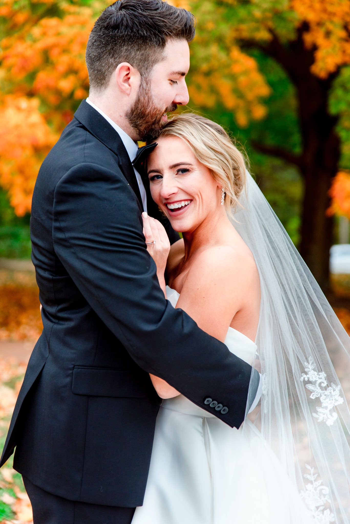 bride with lace veil leaning on grooms chest, who is hugging her in front of orange tree