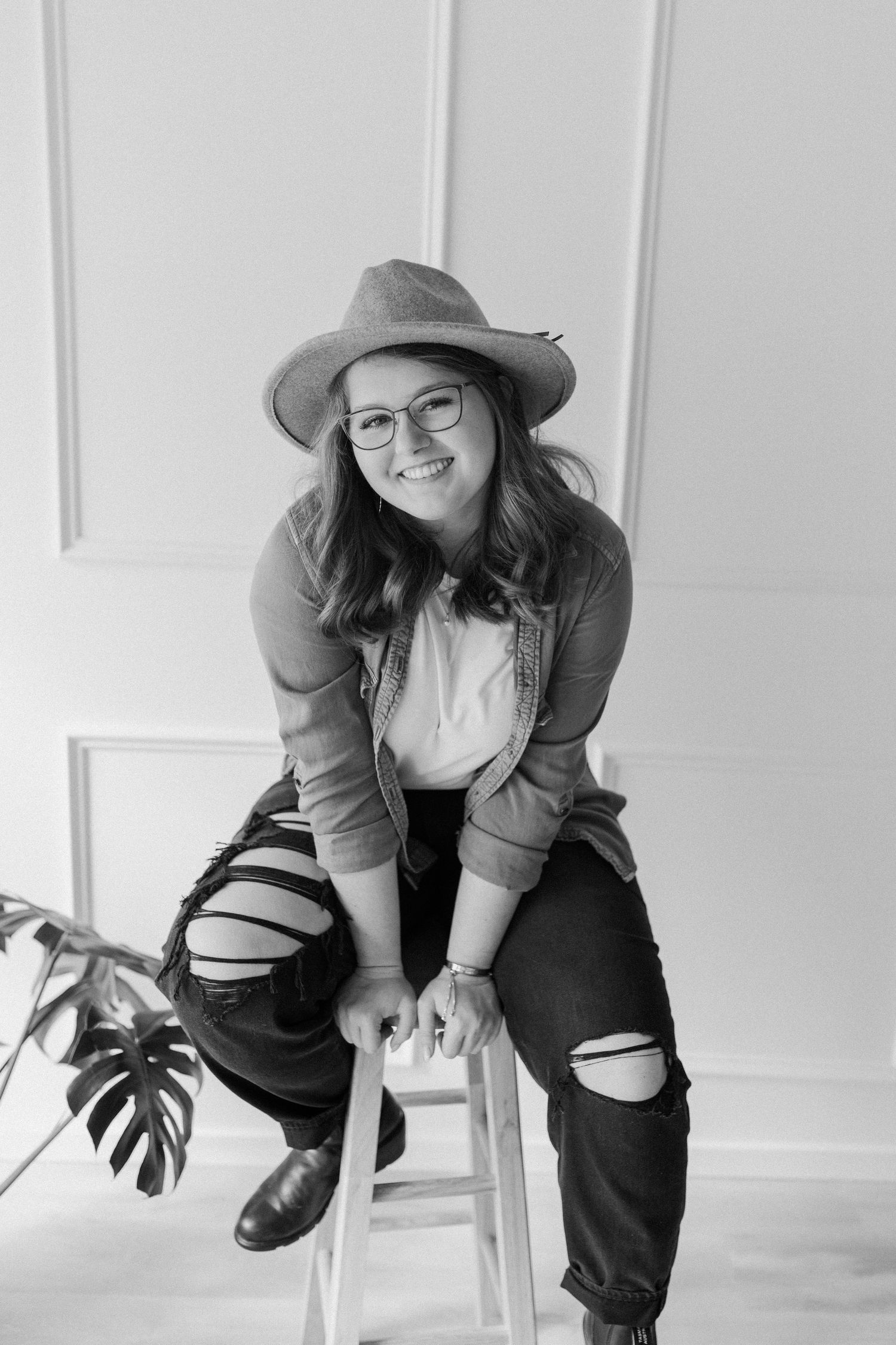 Portrait of a casual model in ripped jeans and hat sitting on stool in bright studio setting.