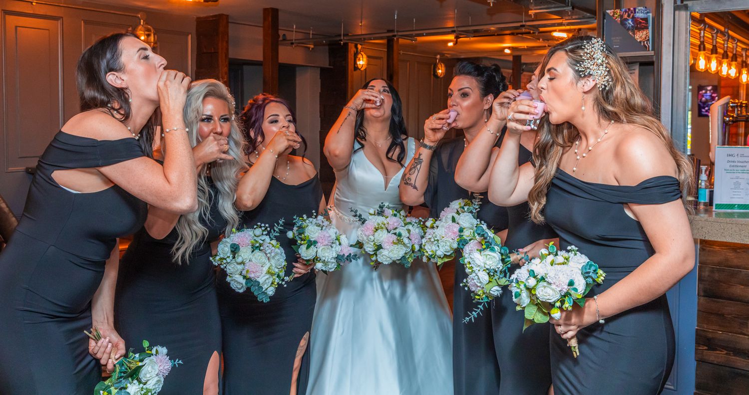 Bridesmaids in navy dresses taking shots with bouquets at elegant indoor reception.