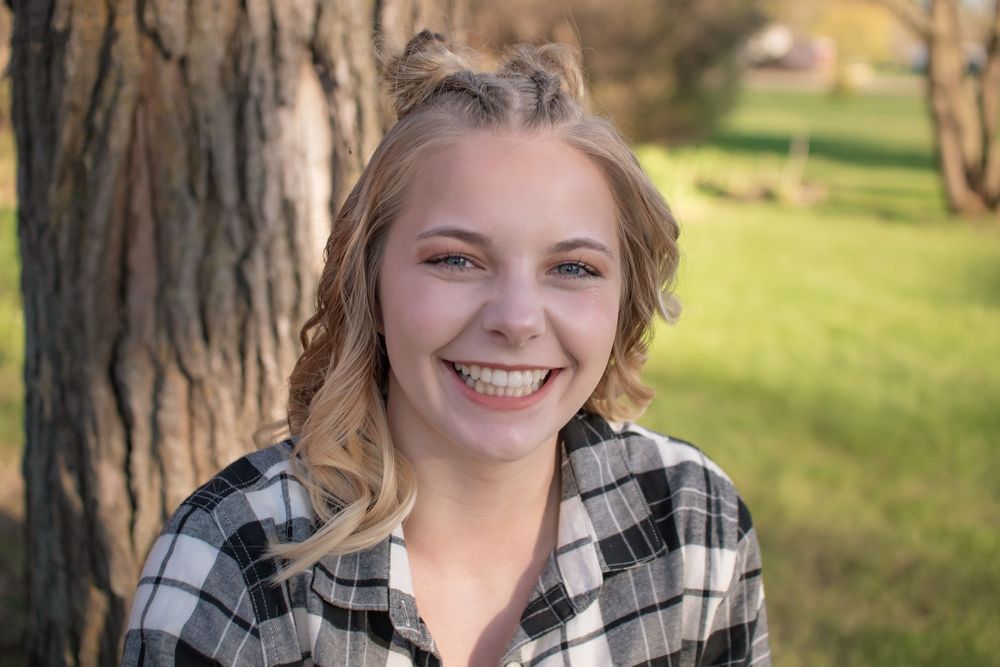 Girl with blond hair wearing black white plaid shirt sitting in front of oak tree