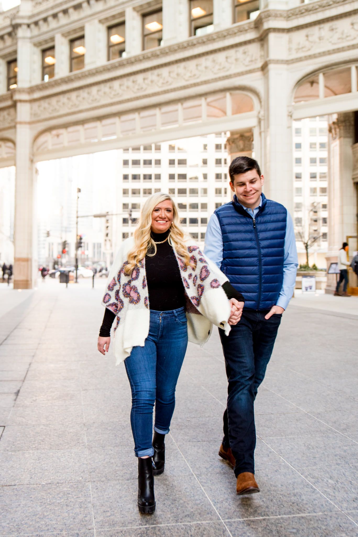 blonde woman in leopard cardigan walking with fiance in blue vest in front for Wrigley Building engagement pictures