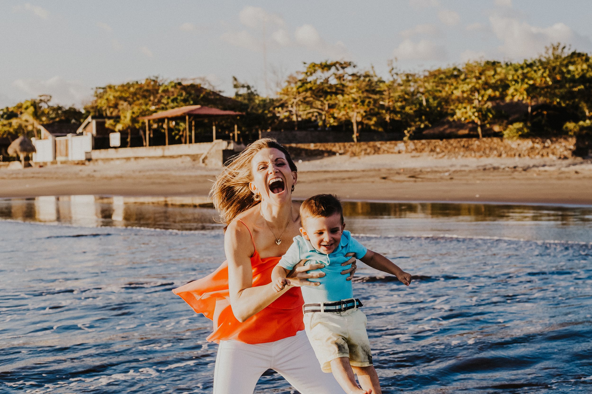 mother laughs while she lifts son into the air over the water on the beach