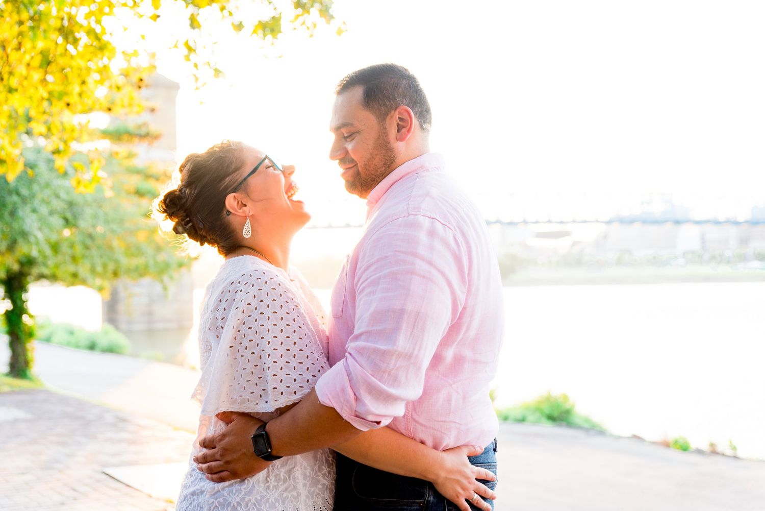 woman in white shirt hugs her fiance in pink shirt and laughs while looking at him under a yellow tree at sunset