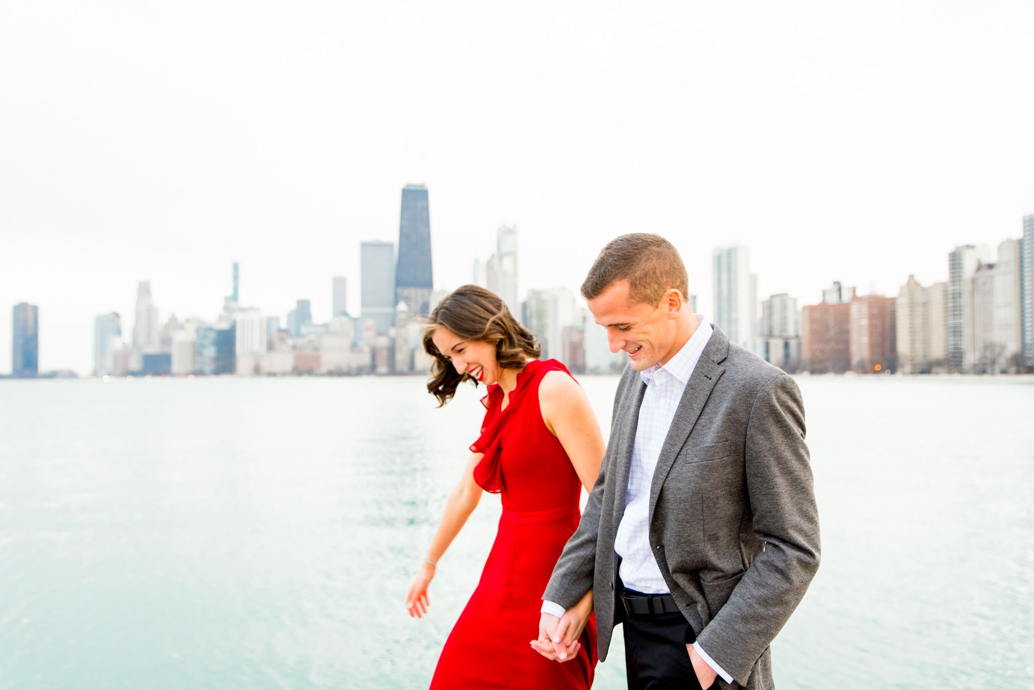 woman in red dress holding hands walking with man in grey suit along North Avenue Beach Chicago engagement pictures