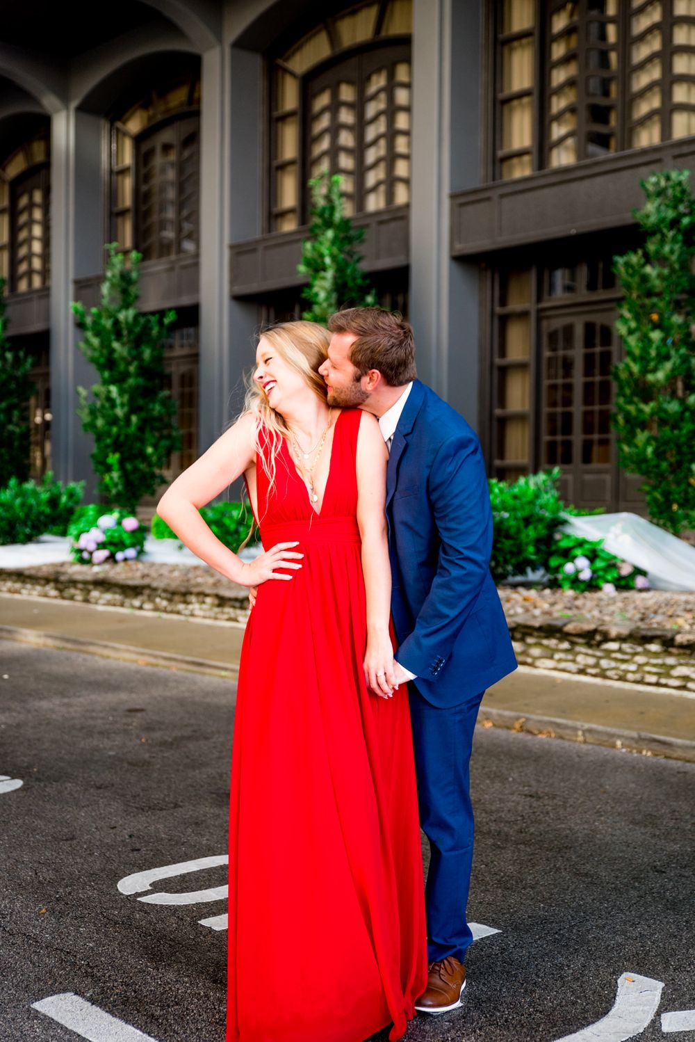 blonde woman in red dress laughing with fiance in navy blue suit tries to kiss her