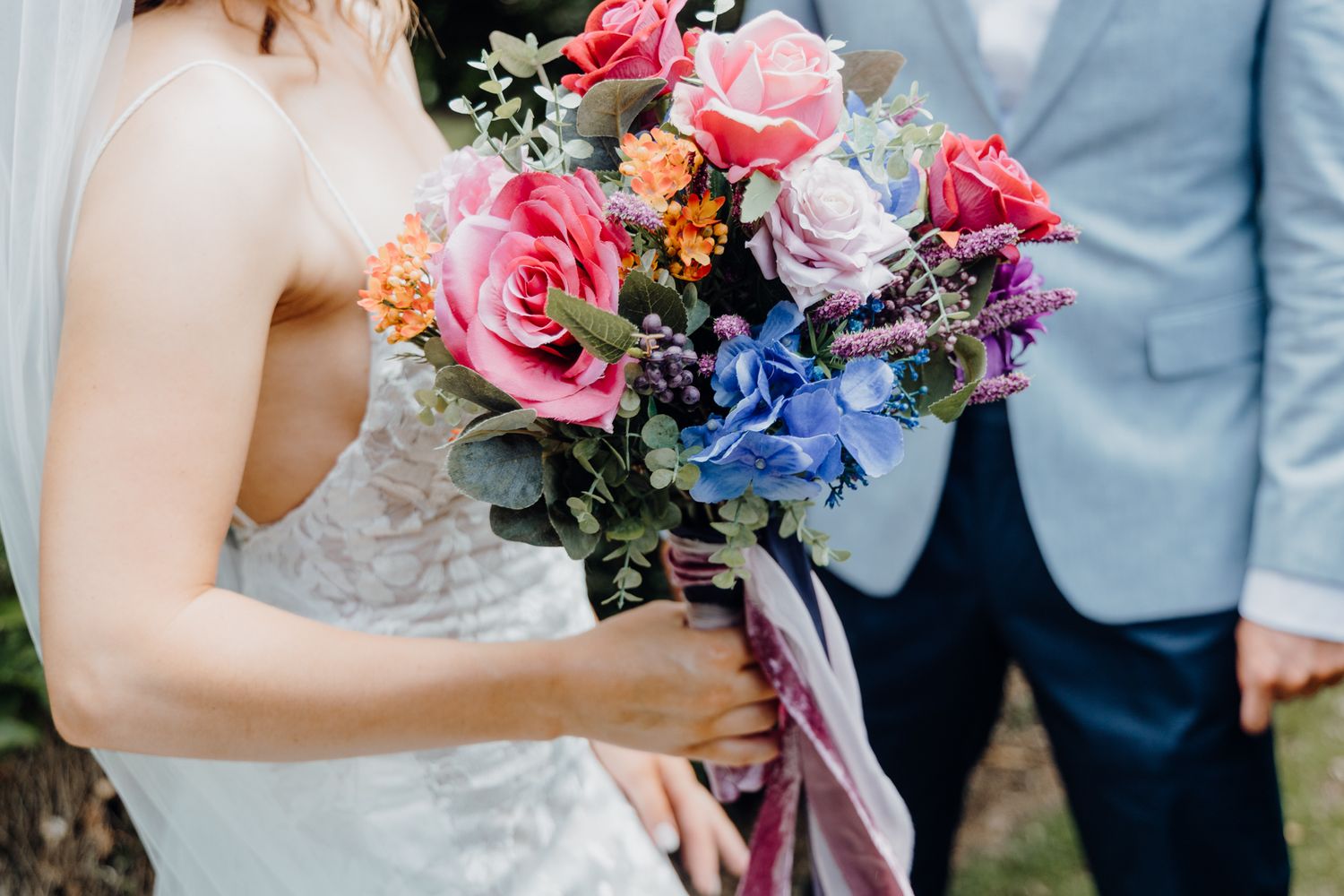 Close-up of a colorful bridal bouquet with pink roses, blue hydrangeas, and purple flowers tied with ribbons.