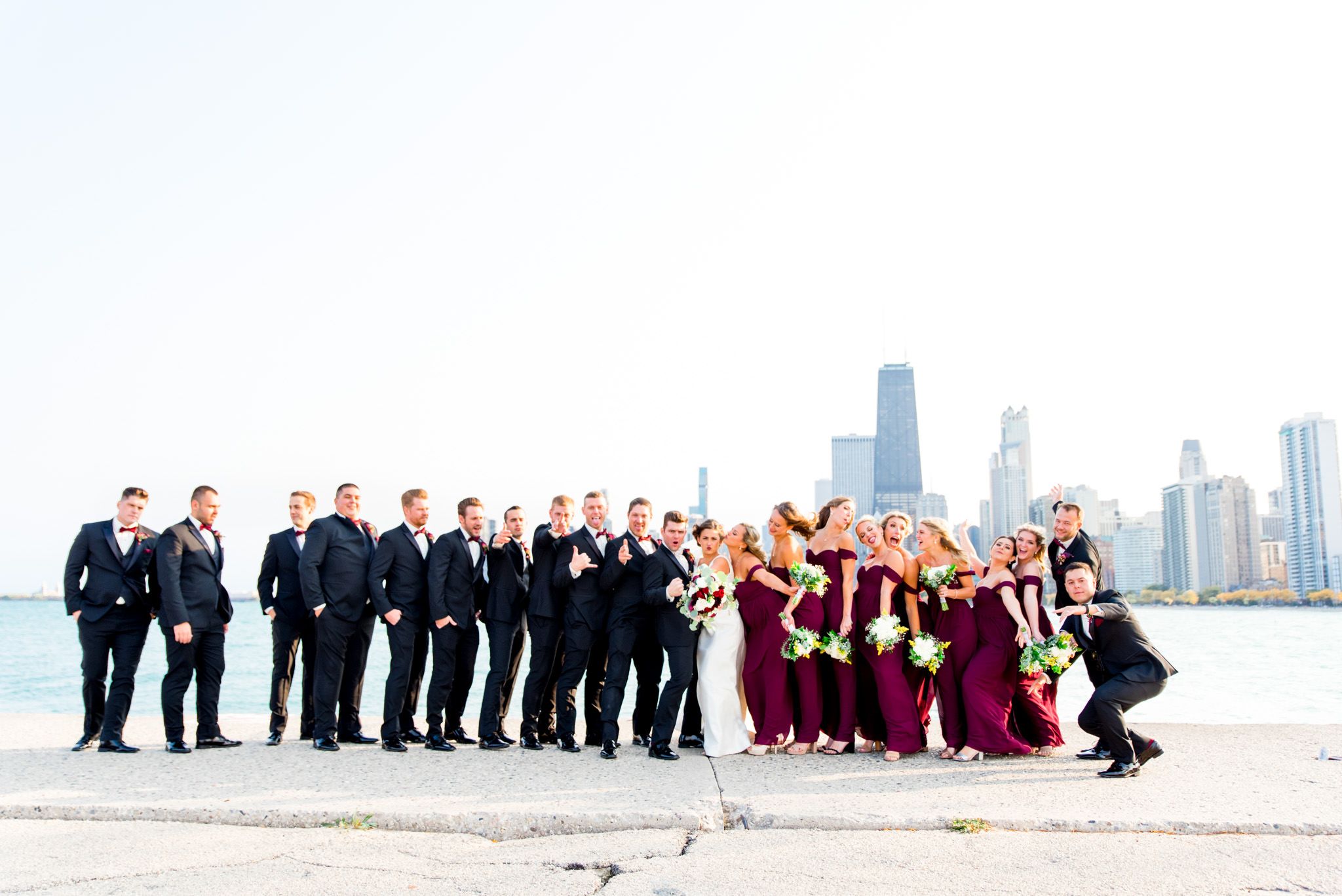 silly wedding party wearing red dresses and black suits strike a pose at north avenue beach pier in chicago