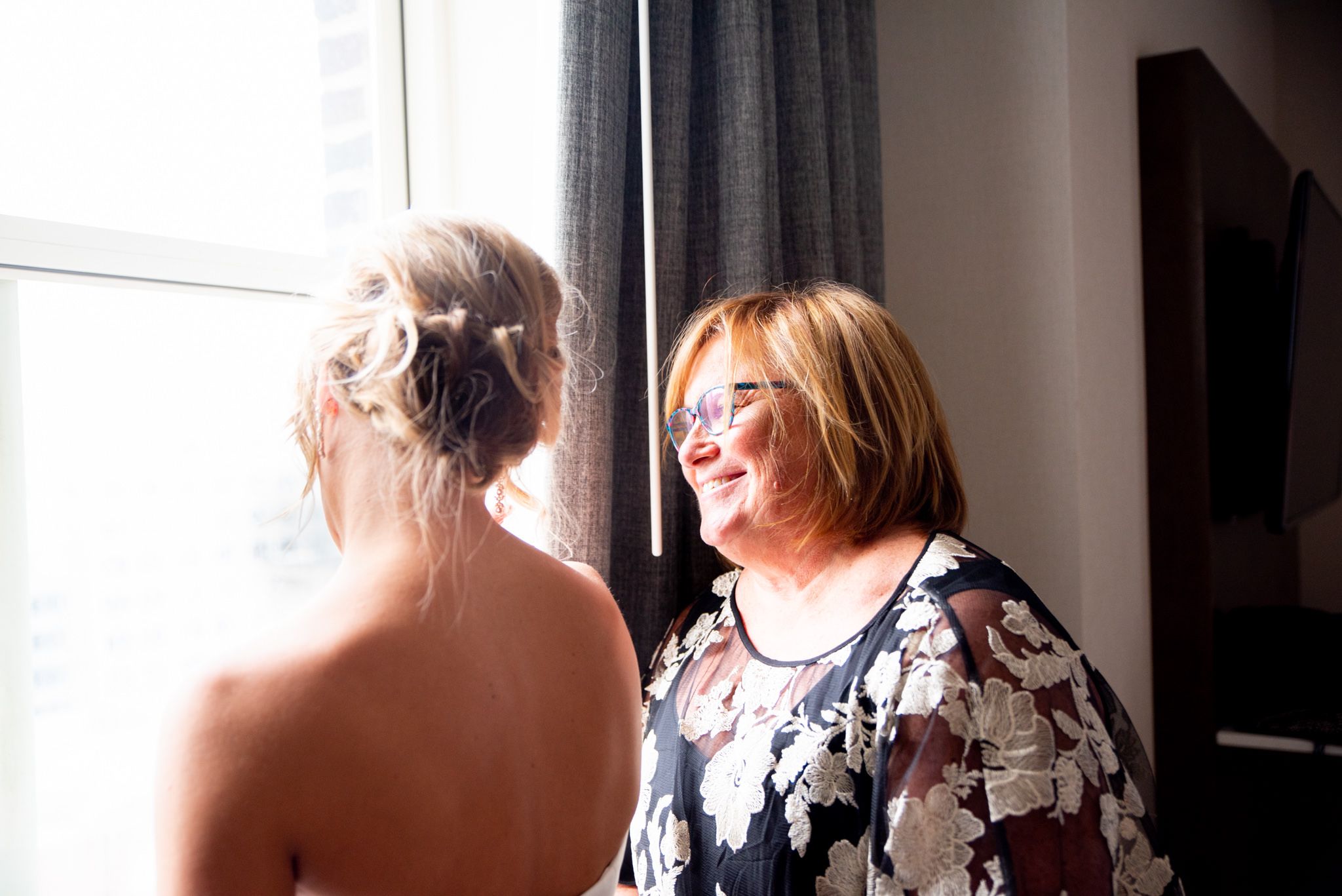 mother of the bride smiling at her daughter while getting ready next to a window