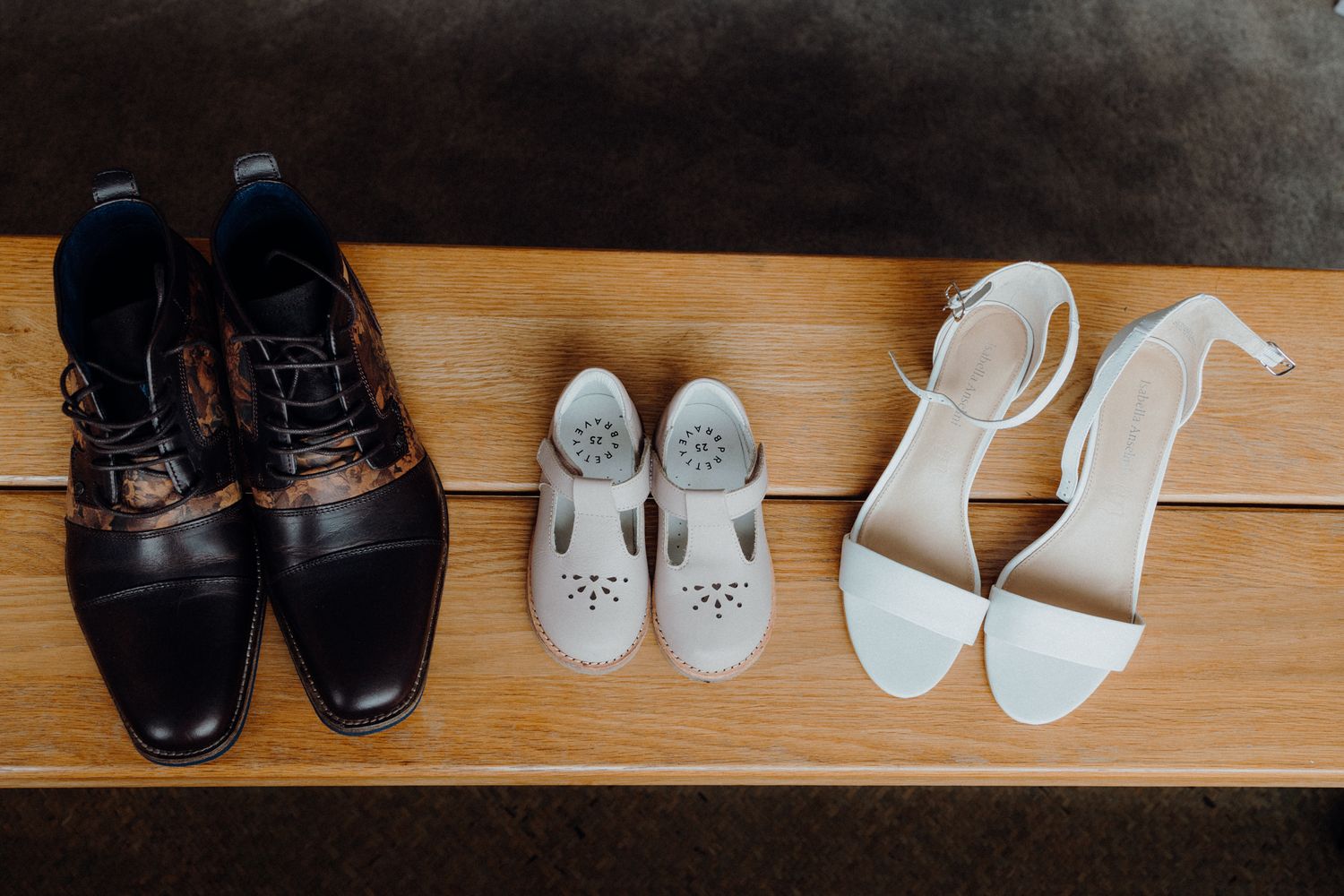 Three pairs of wedding shoes lined up on wooden bench including black dress shoes, white flats, and white heels.