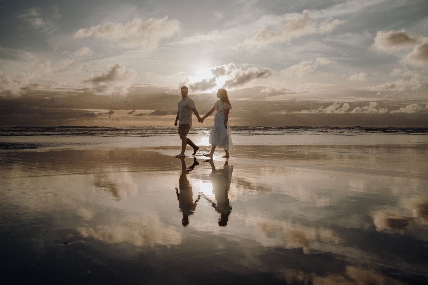 Couple holding hands with perfect reflection in wet beach sand at sunset.