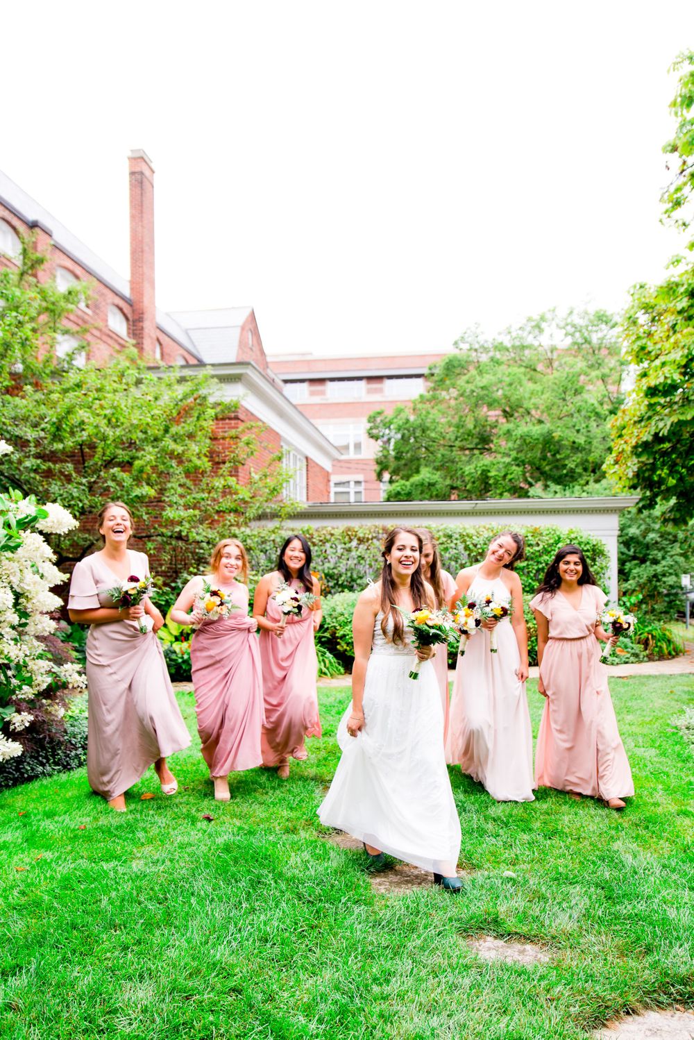 bride and bridesmaids in blush hold bouquets and walk through garden laughing and smiling