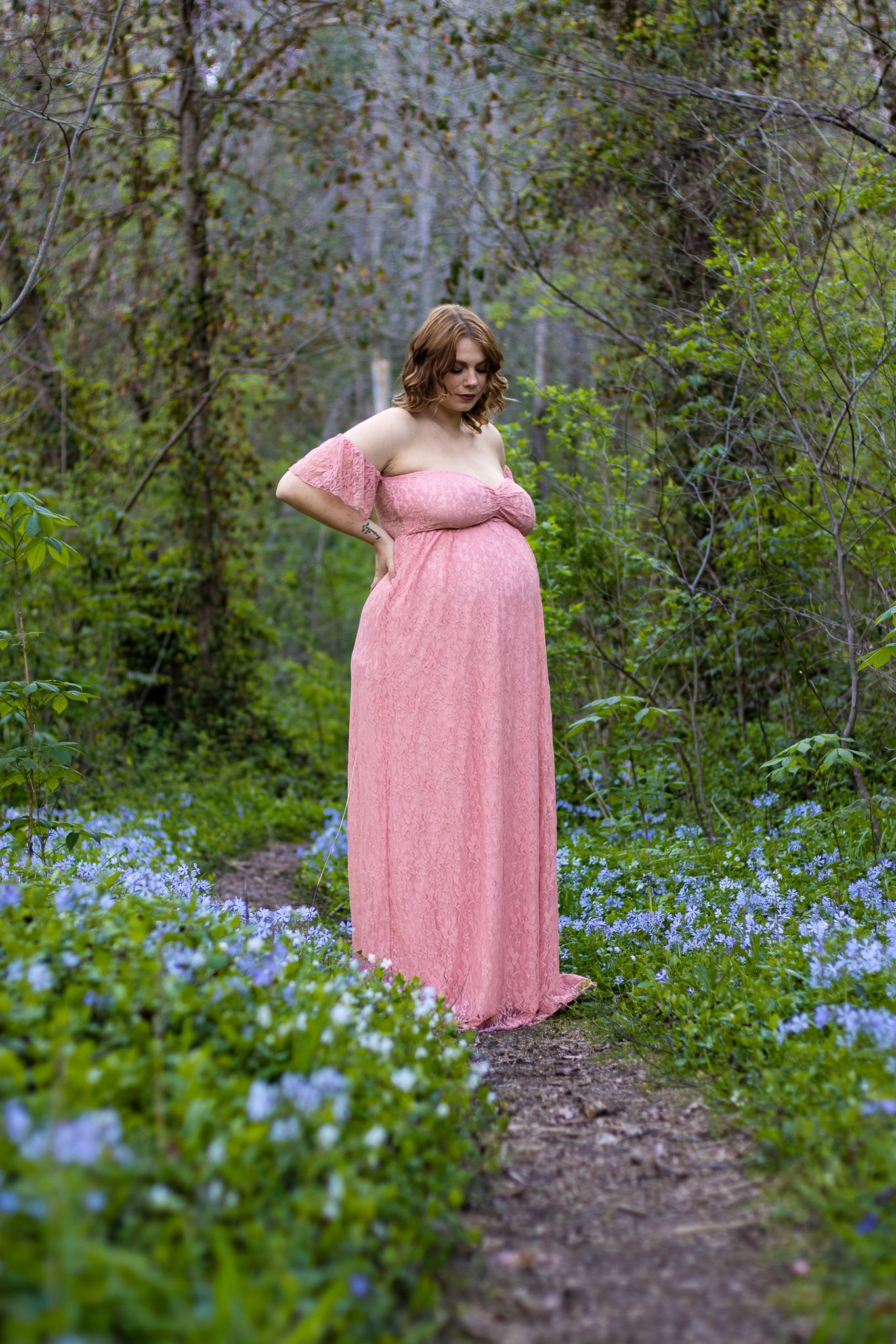 A pink floral maternity gown worn in a wooded setting with blue wildflowers blooming along the path.
