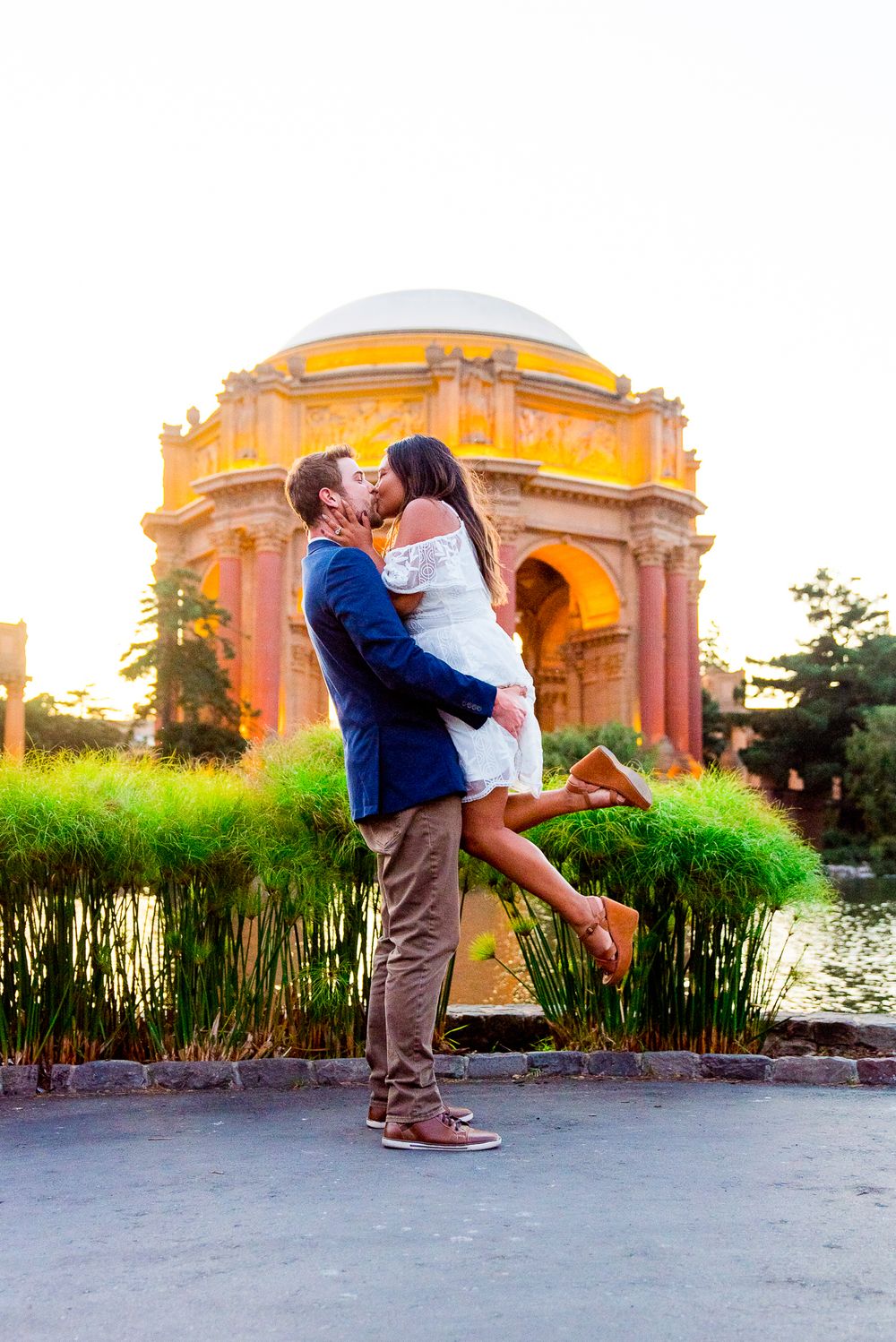 man in blue suit picking up woman in white dress kissing in front of Palace of Fine Arts in the summer