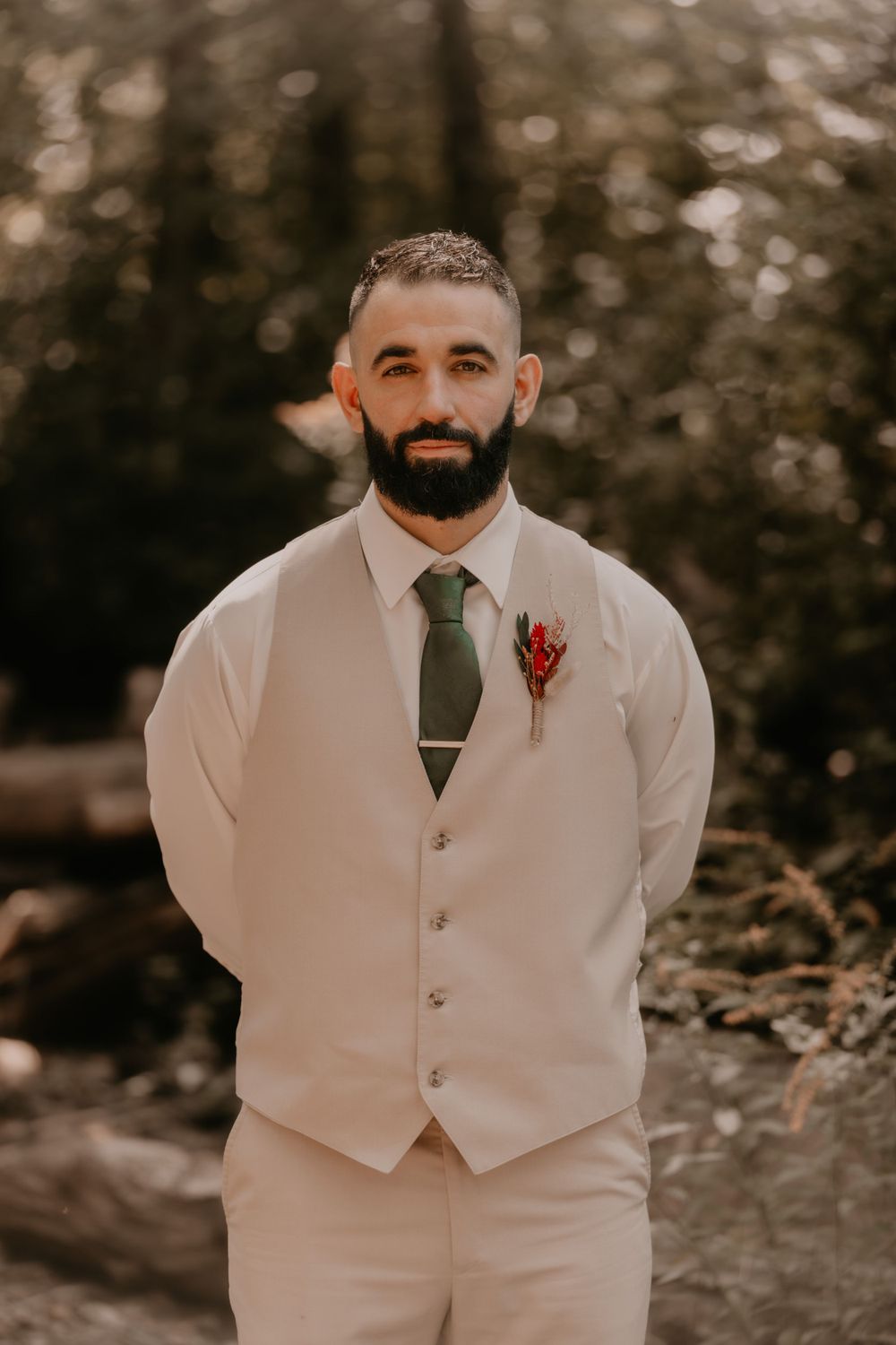 Groom in light beige vest and green tie smiles at the camera in a forest setting.