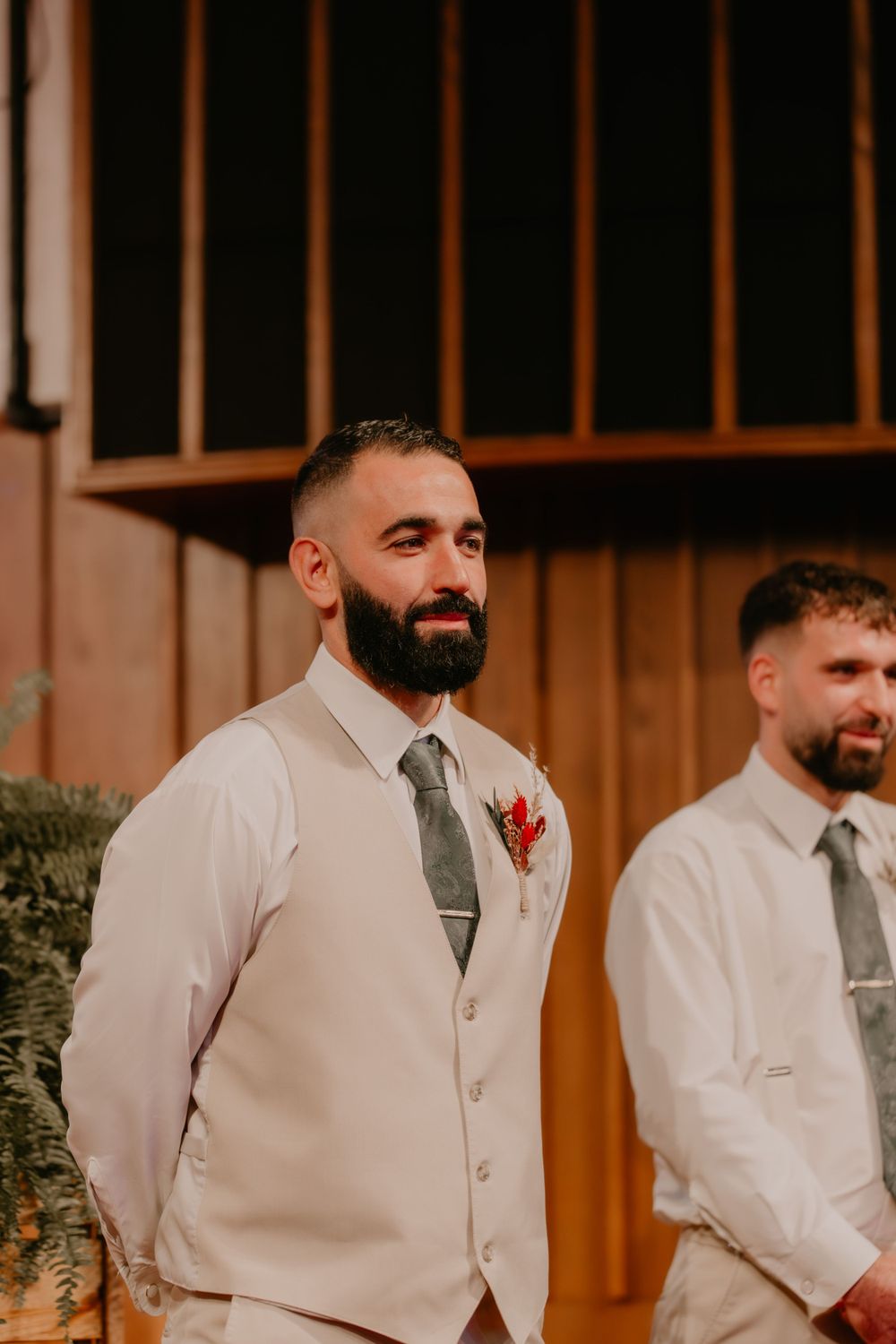 Two groomsmen in light beige vests and gray ties stand together smiling.