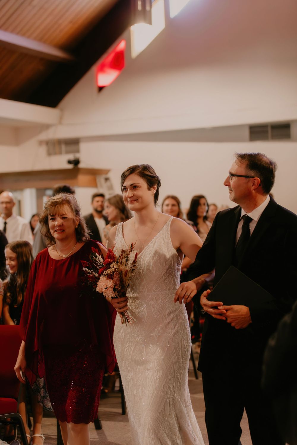 A bride walks down the aisle in a fitted lace gown accompanied by family members.