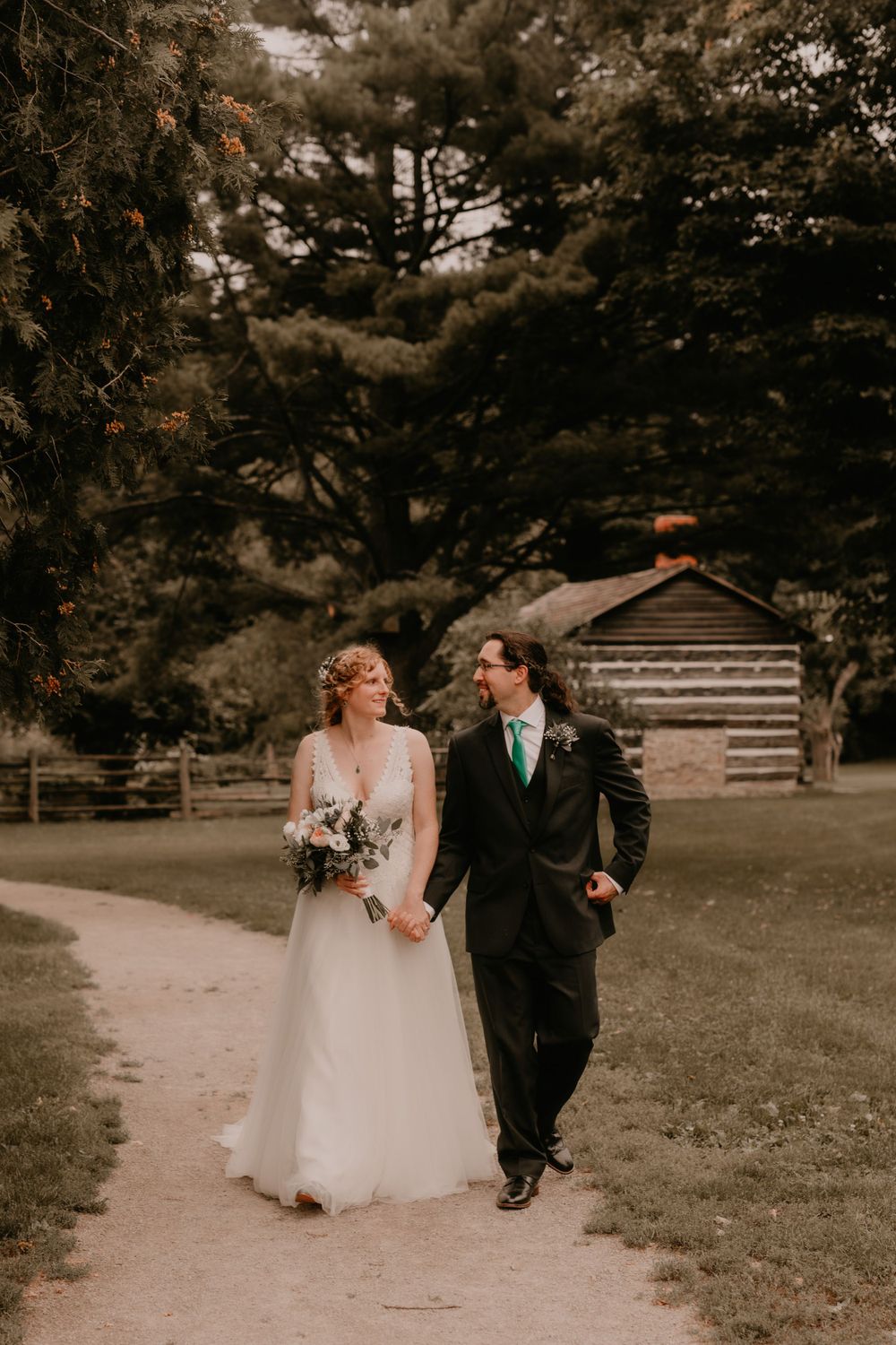 Bride and groom walking hand in hand on path with rustic cabin in background.