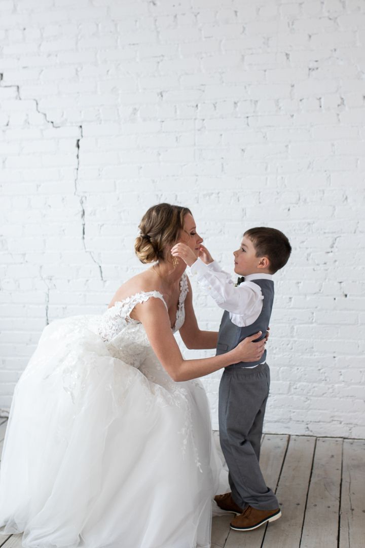 Bride and ring bearer about to embrace into a hug. white brick wall behind them. boy in grey suit.