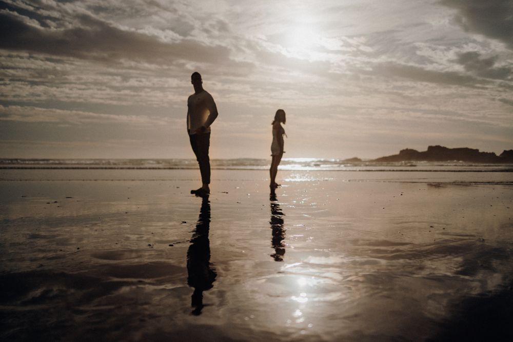 Two silhouettes stand reflected in wet sand during a dramatic beach sunset.