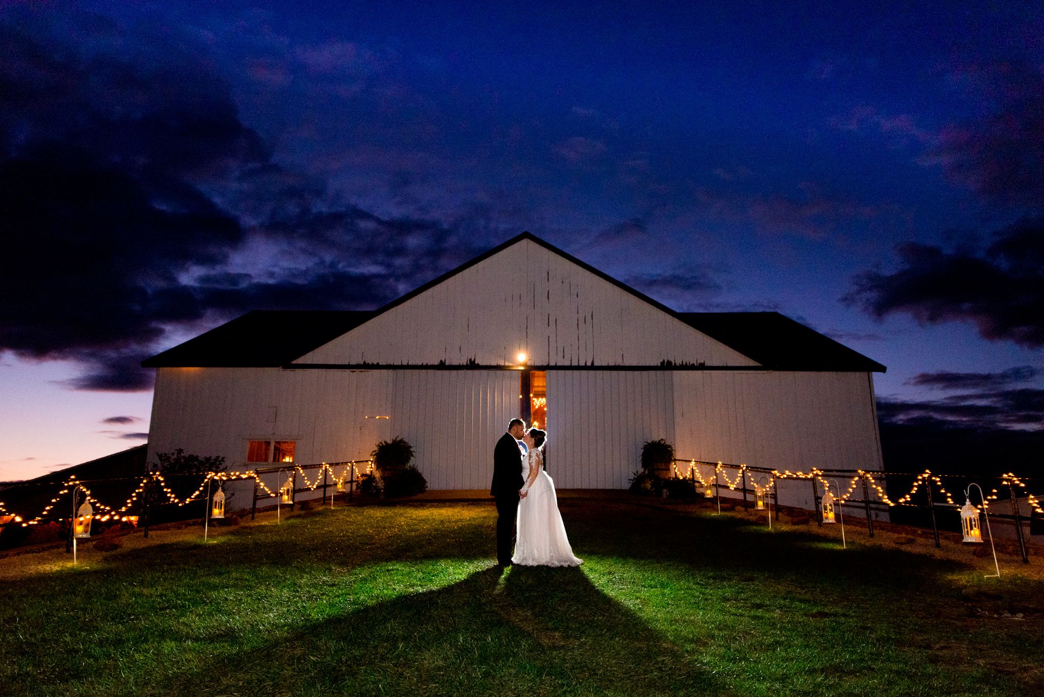 bride and groom kissing in front of white barn at night with dark blue sky and clouds
