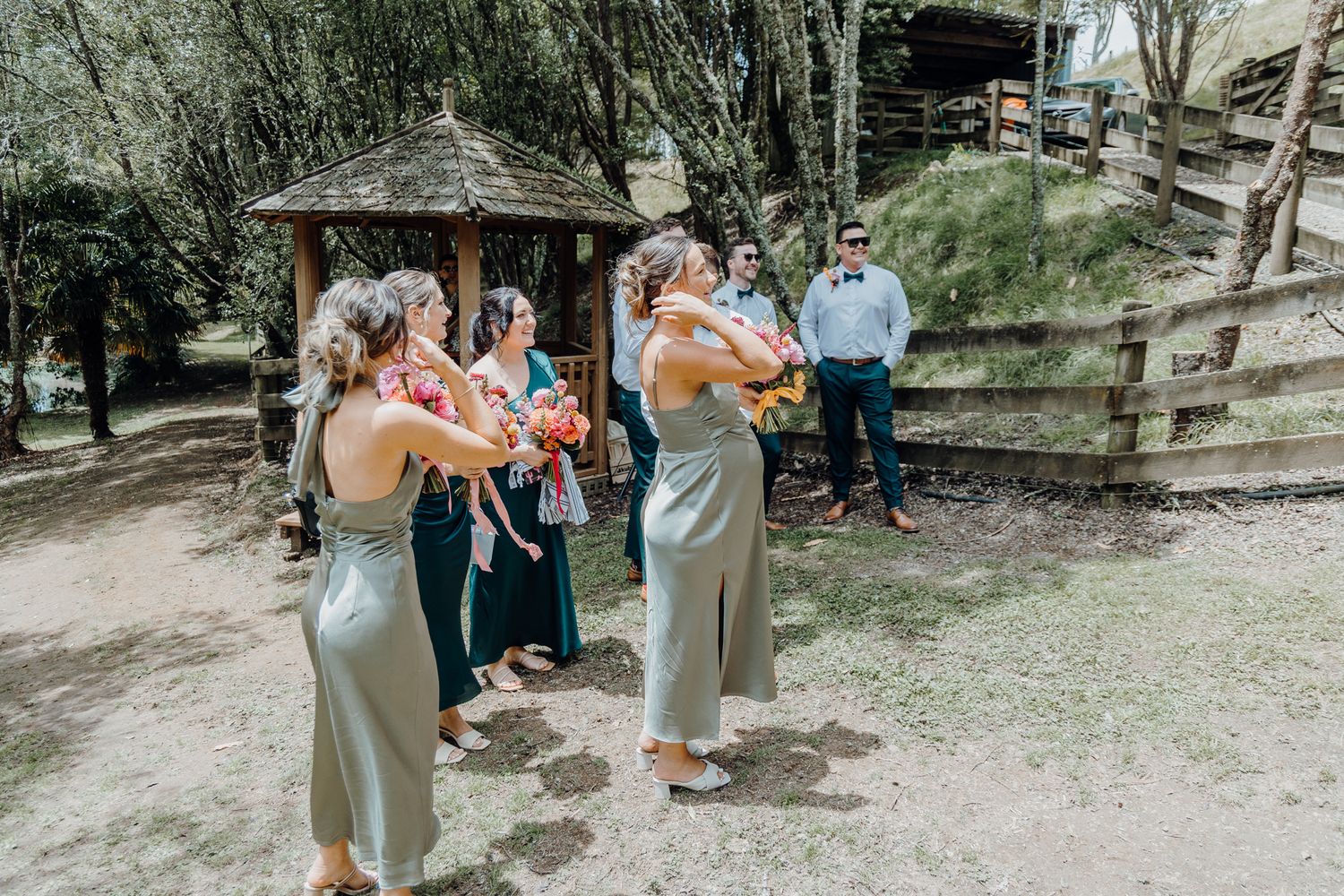 Wedding party gathers under a rustic gazebo holding colorful floral bouquets.