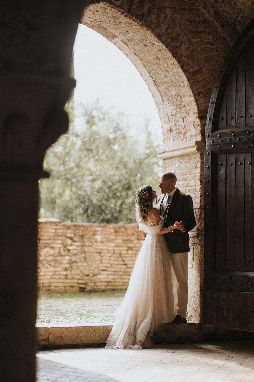 Bride and groom embrace under rustic stone arches with natural light streaming in during romantic wedding photo session.