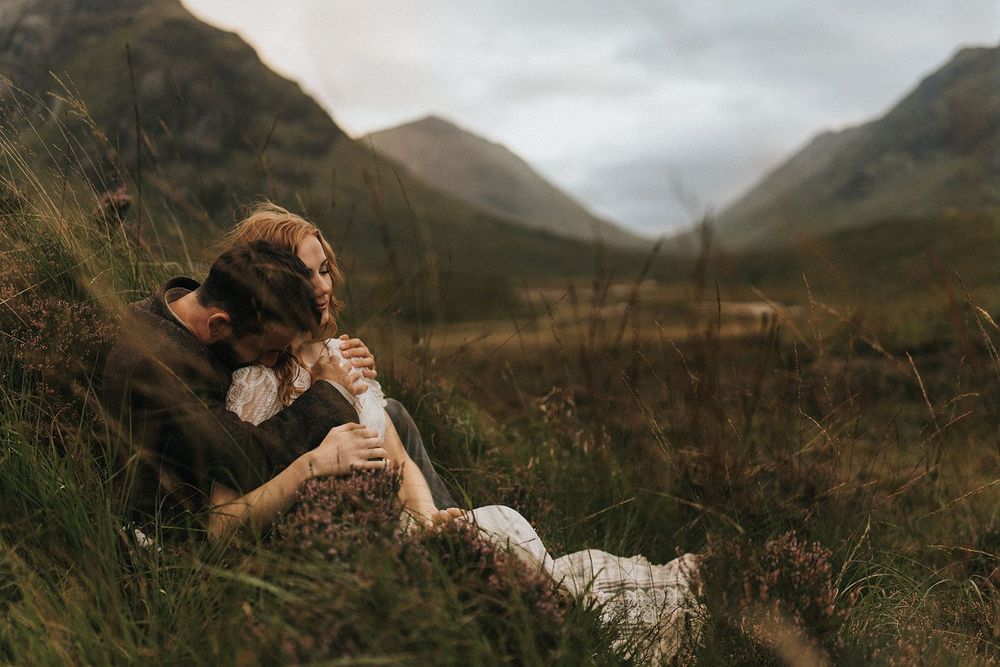 A couple rest together on a grassy hillside in a dramatic Scottish mountain landscape with moody clouds.
