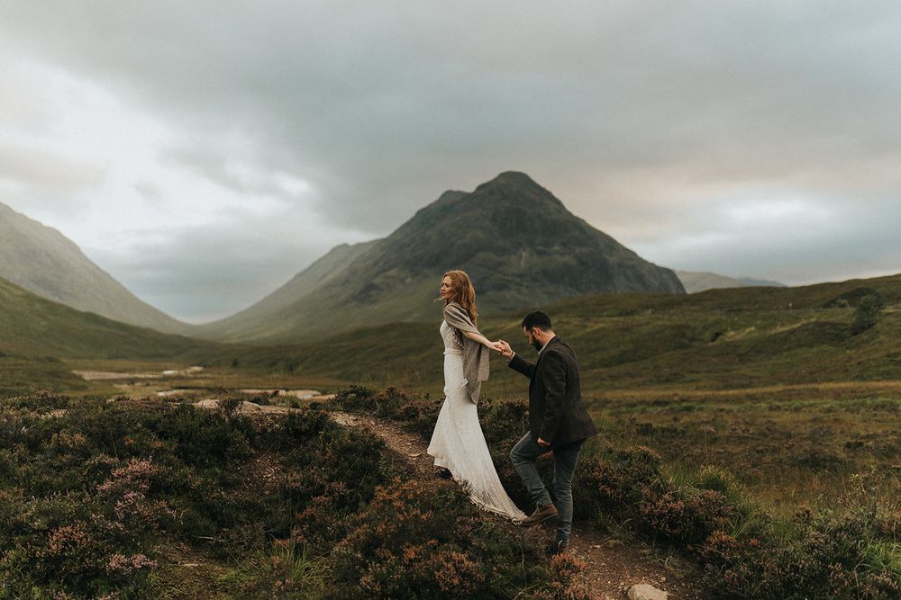 A couple stands in the rugged Scottish Highlands with dramatic mountains and moody skies in the background.