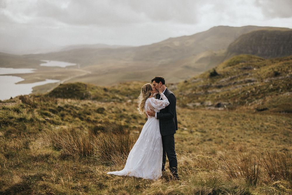 A couple in wedding attire embrace on a scenic hillside overlooking misty lakes and mountains in Scotland.