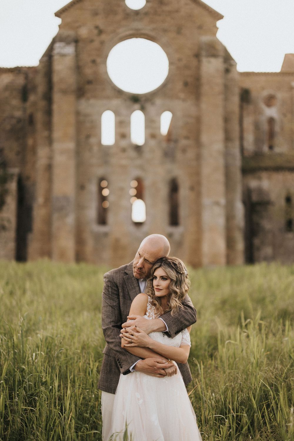 A romantic moment between a couple embracing in tall grass by a rustic stone church ruin.