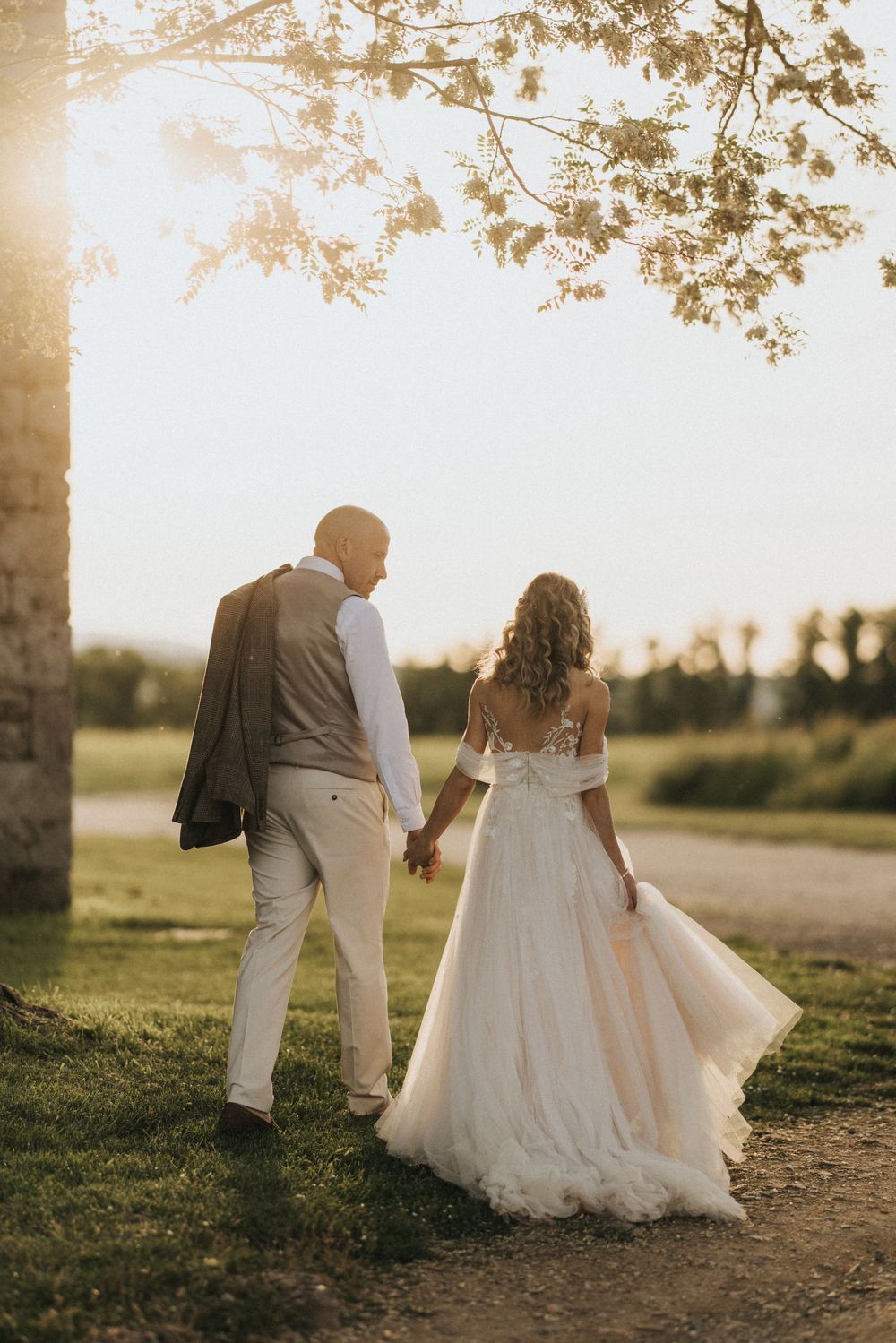 A romantic wedding photo shows a couple walking hand in hand at sunset in a grassy field.