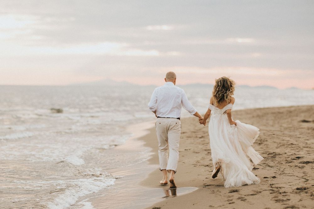 A couple in white wedding attire holds hands while walking along the beach during sunset.