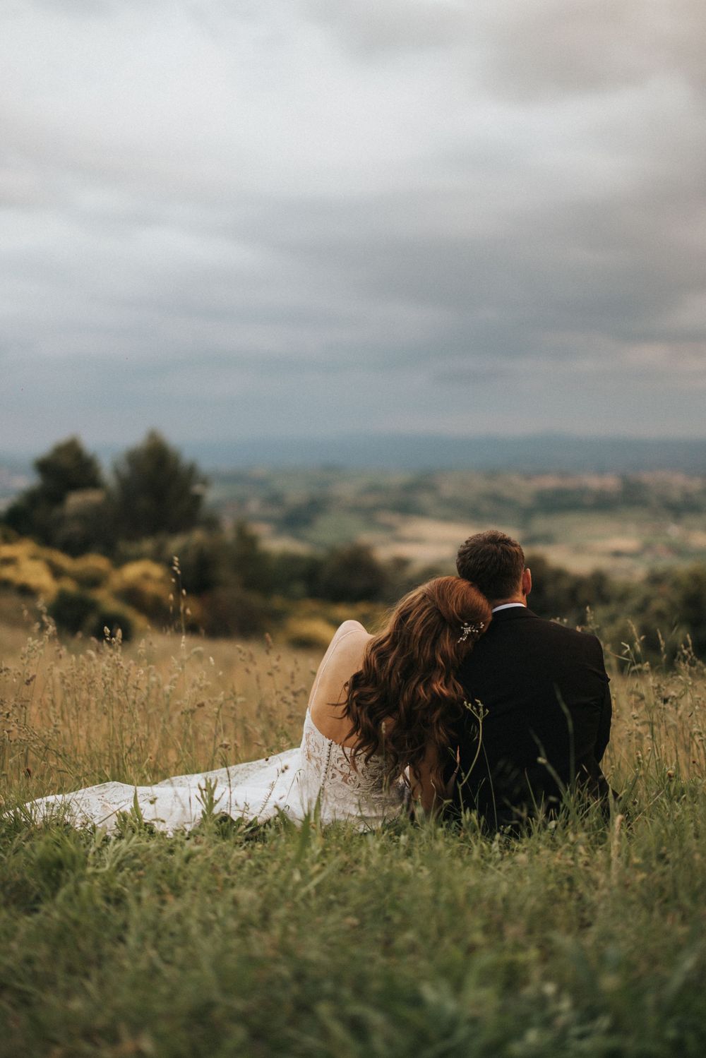 A couple lies together in a serene golden meadow beneath cloudy skies in Italy.