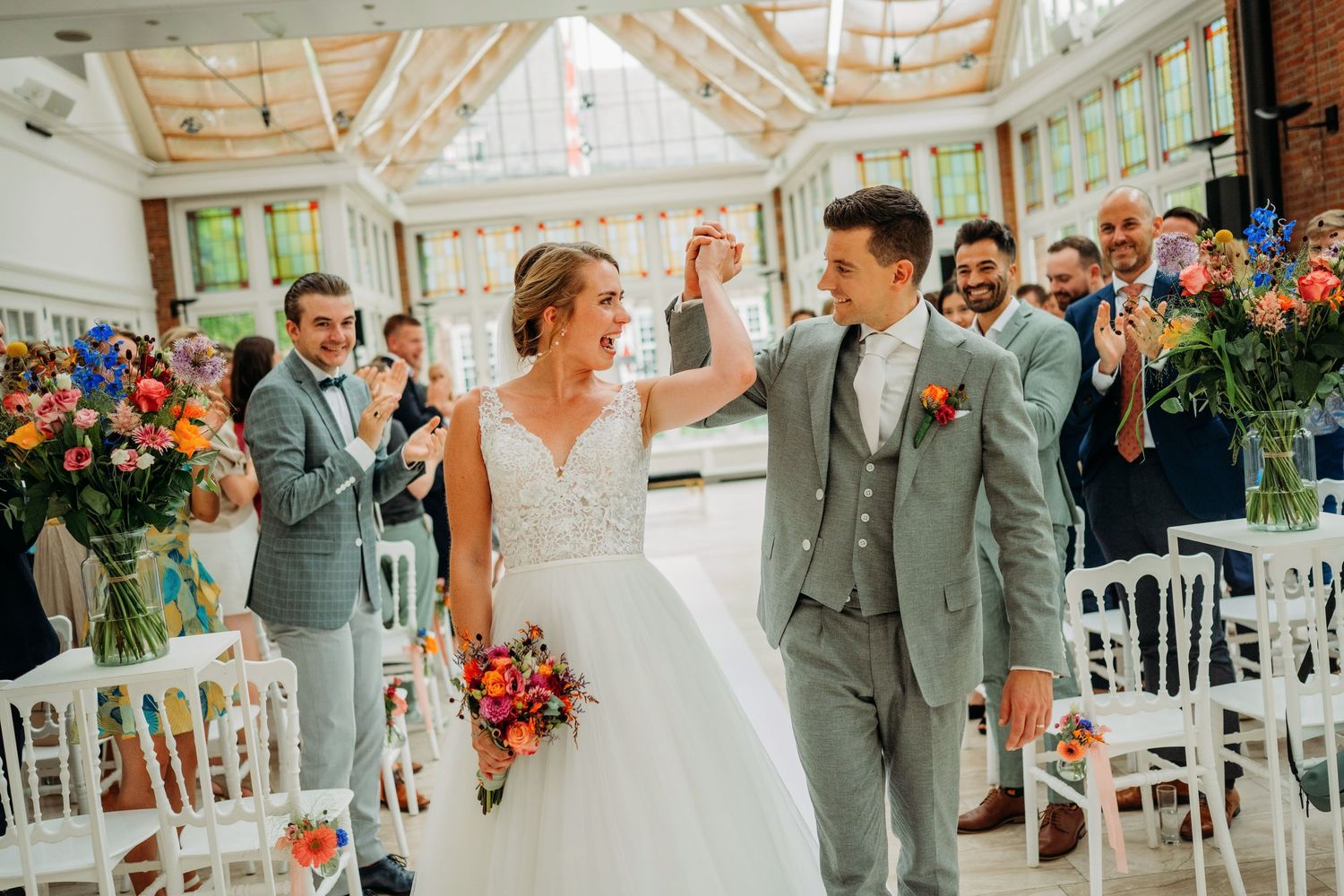 Two people in grey and white attire celebrate with guests inside an elegant conservatory filled with colorful flowers.