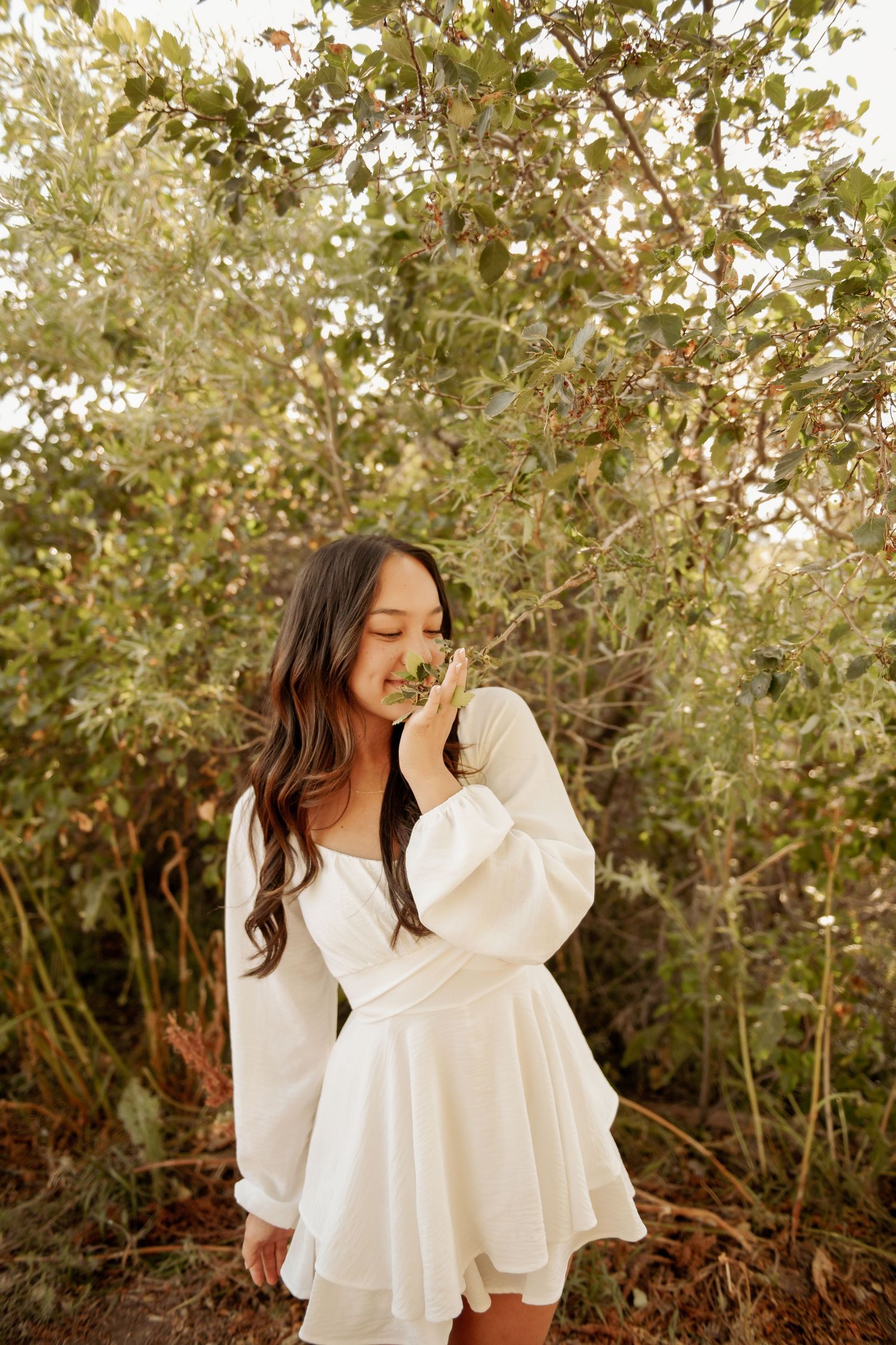 A woman wearing a flowy white dress with puffy sleeves stands gracefully in front of lush green foliage during golden hour.