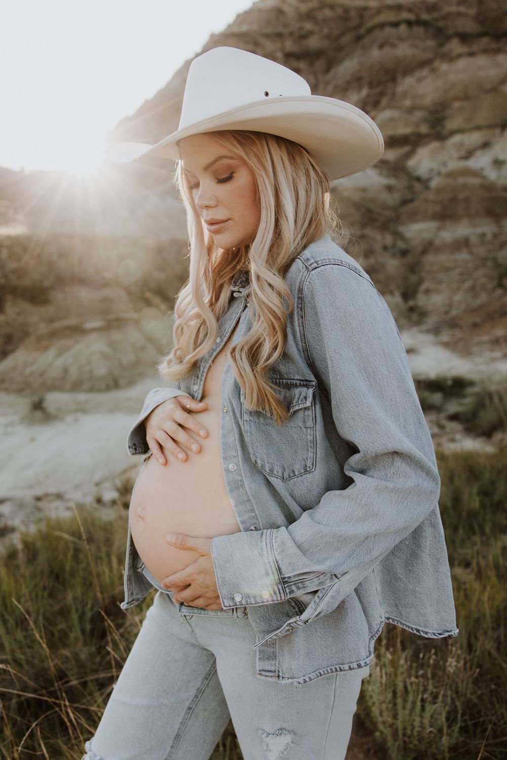 A soft, ethereal maternity photo shoot on a cliffside against golden lighting shows a woman in denim and cowboy hat.